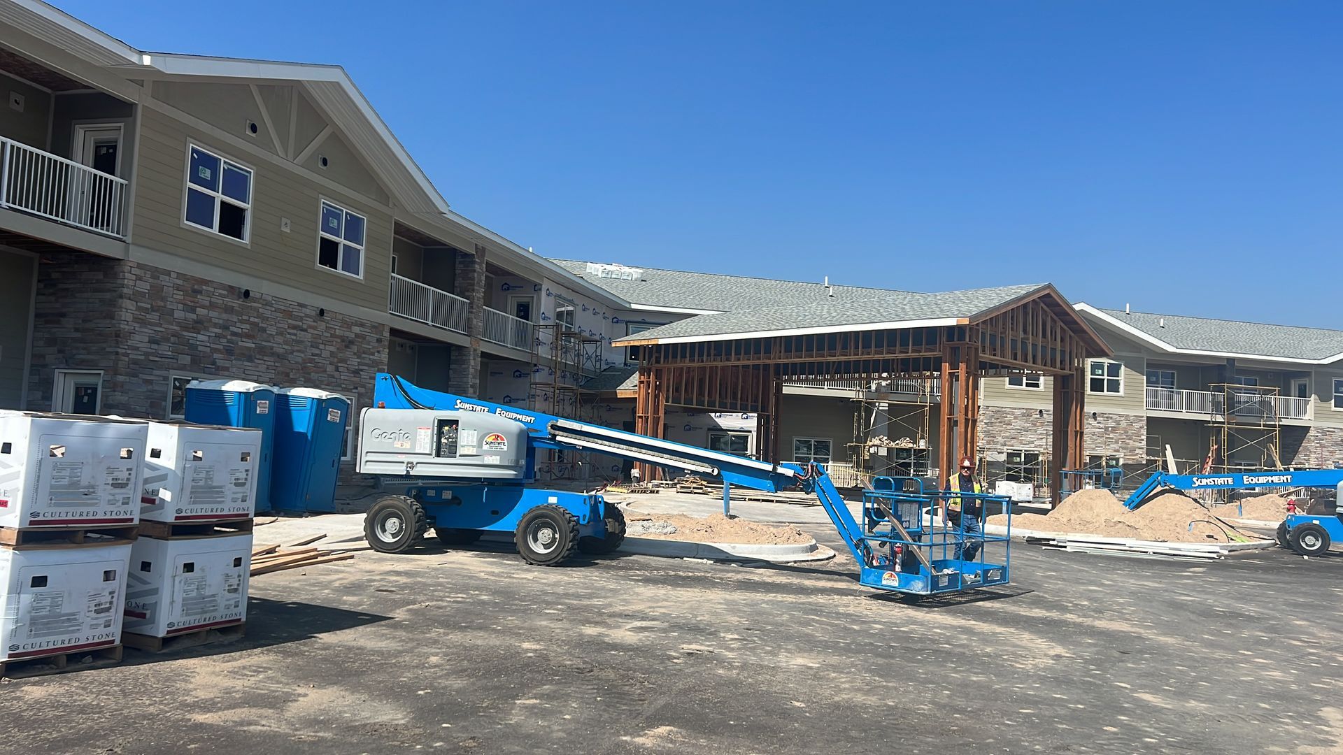 Construction site with a partially built beige and stone building, blue lifts, and materials. Sunny, clear day.