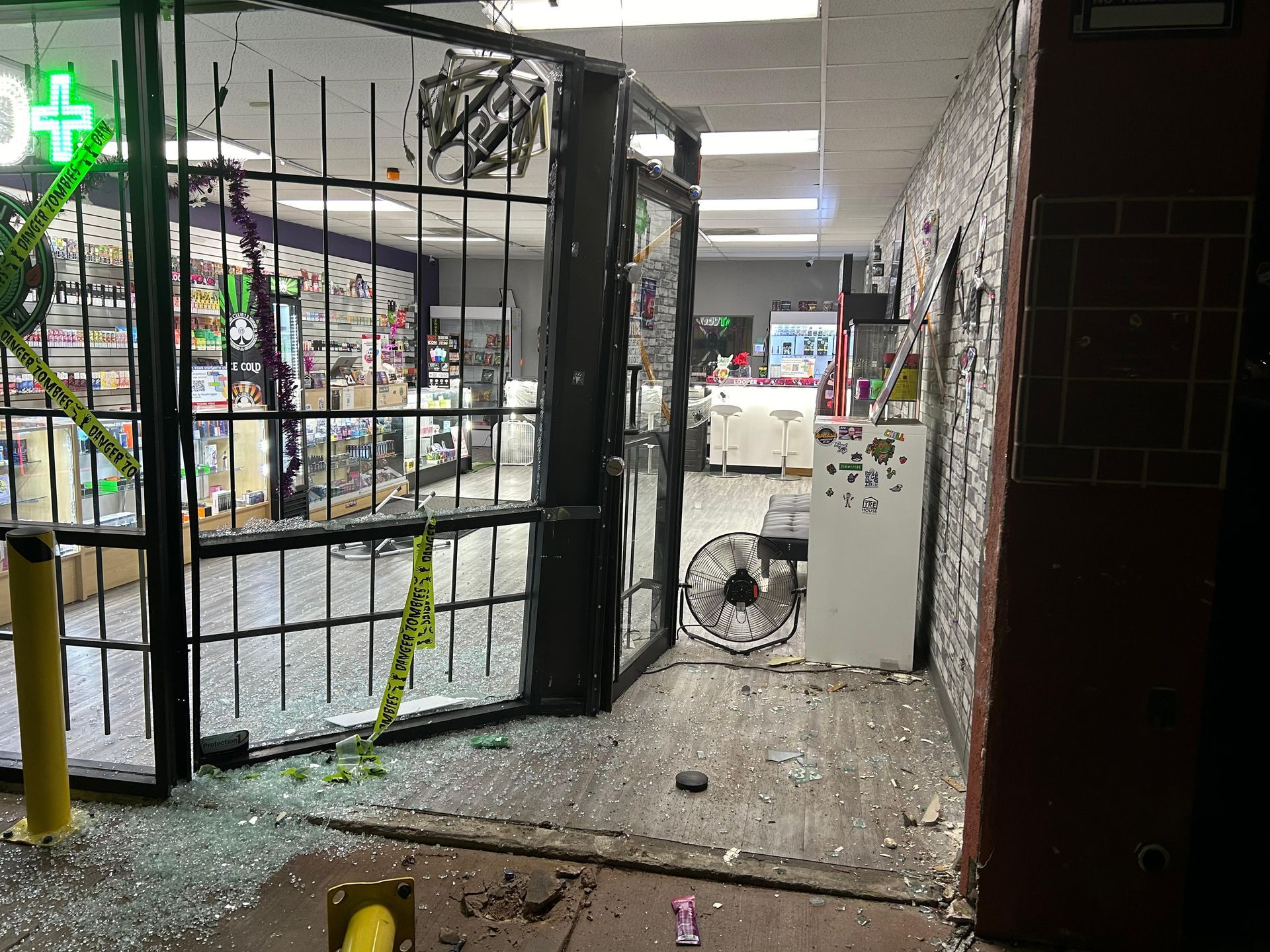Shattered storefront entrance with broken glass and security gate; inside retail store visible.