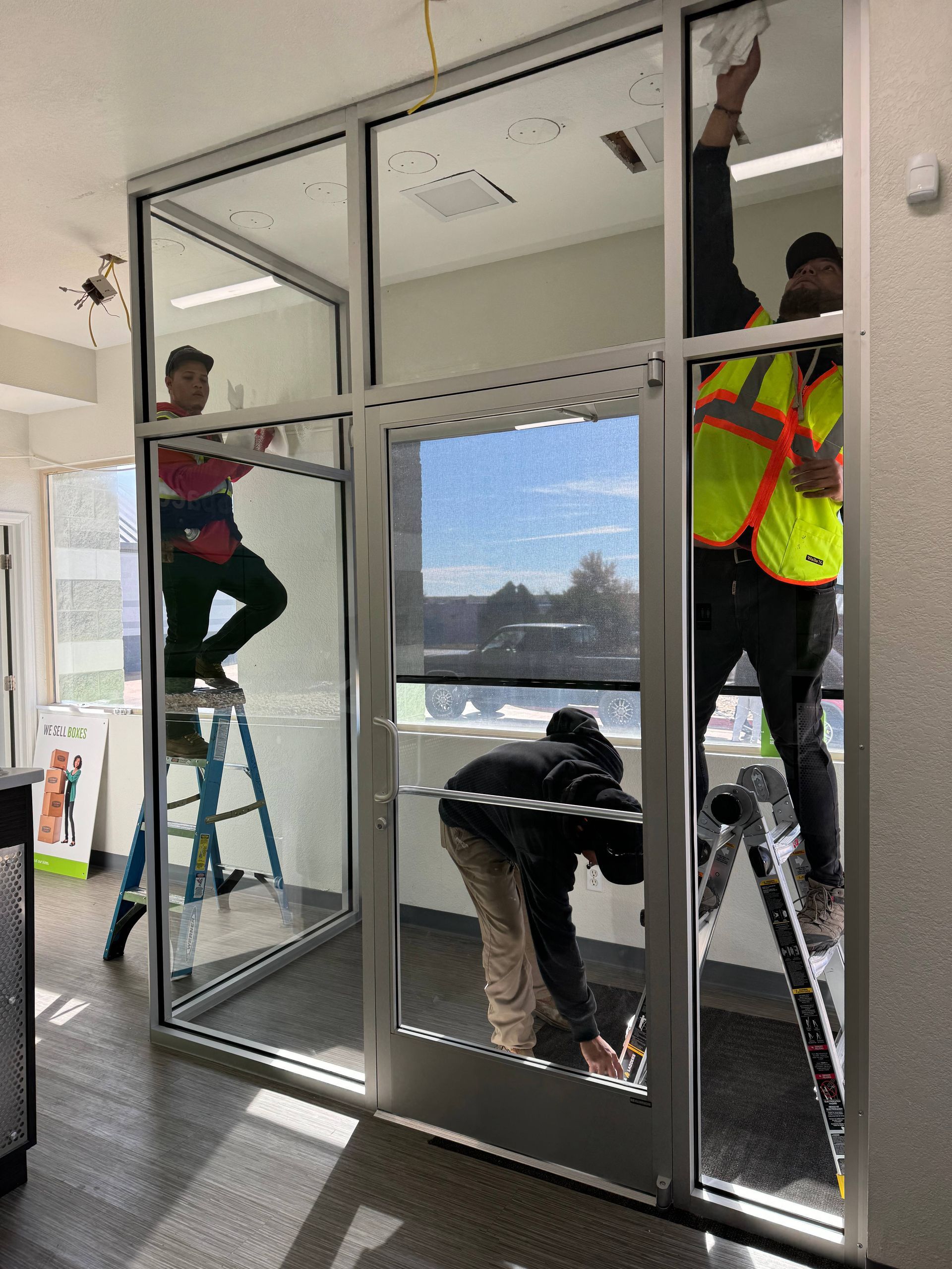 Three workers installing glass door in building. One on ladder, others working on frame.