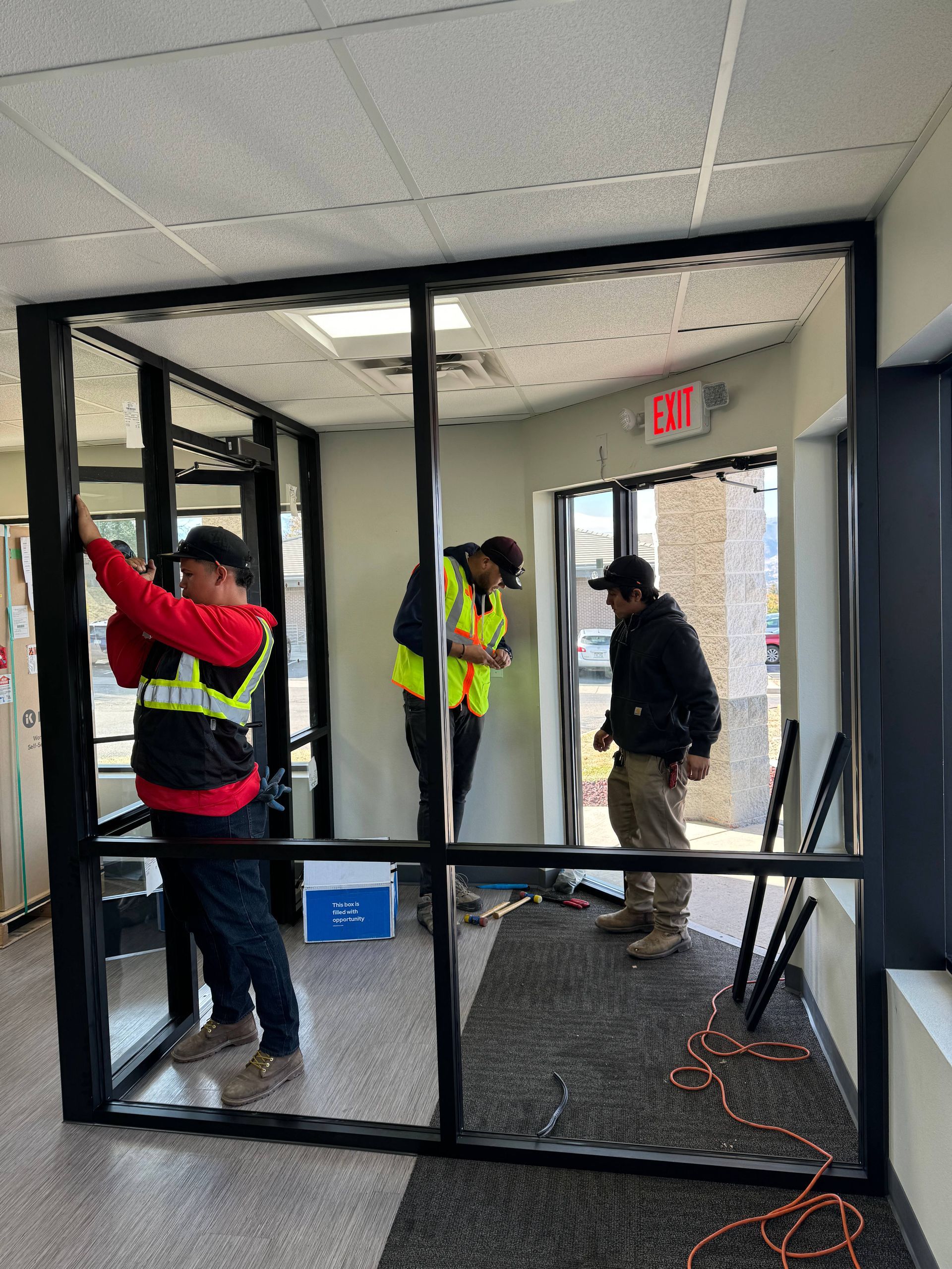 Three workers installing glass panels in a commercial interior. Black frames, safety vests.