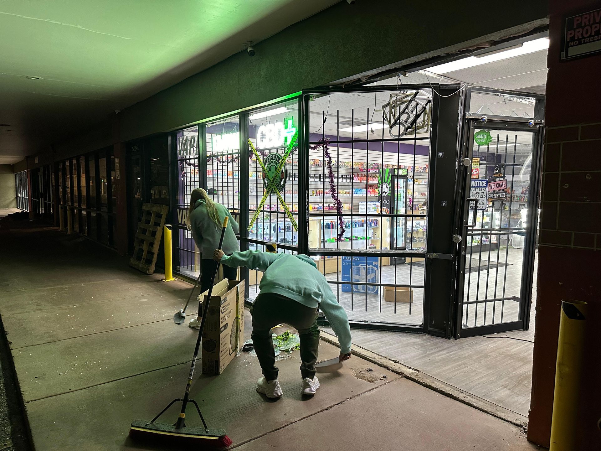 Two people sweeping debris from a store entrance with damaged security gates. Indoor hallway setting.