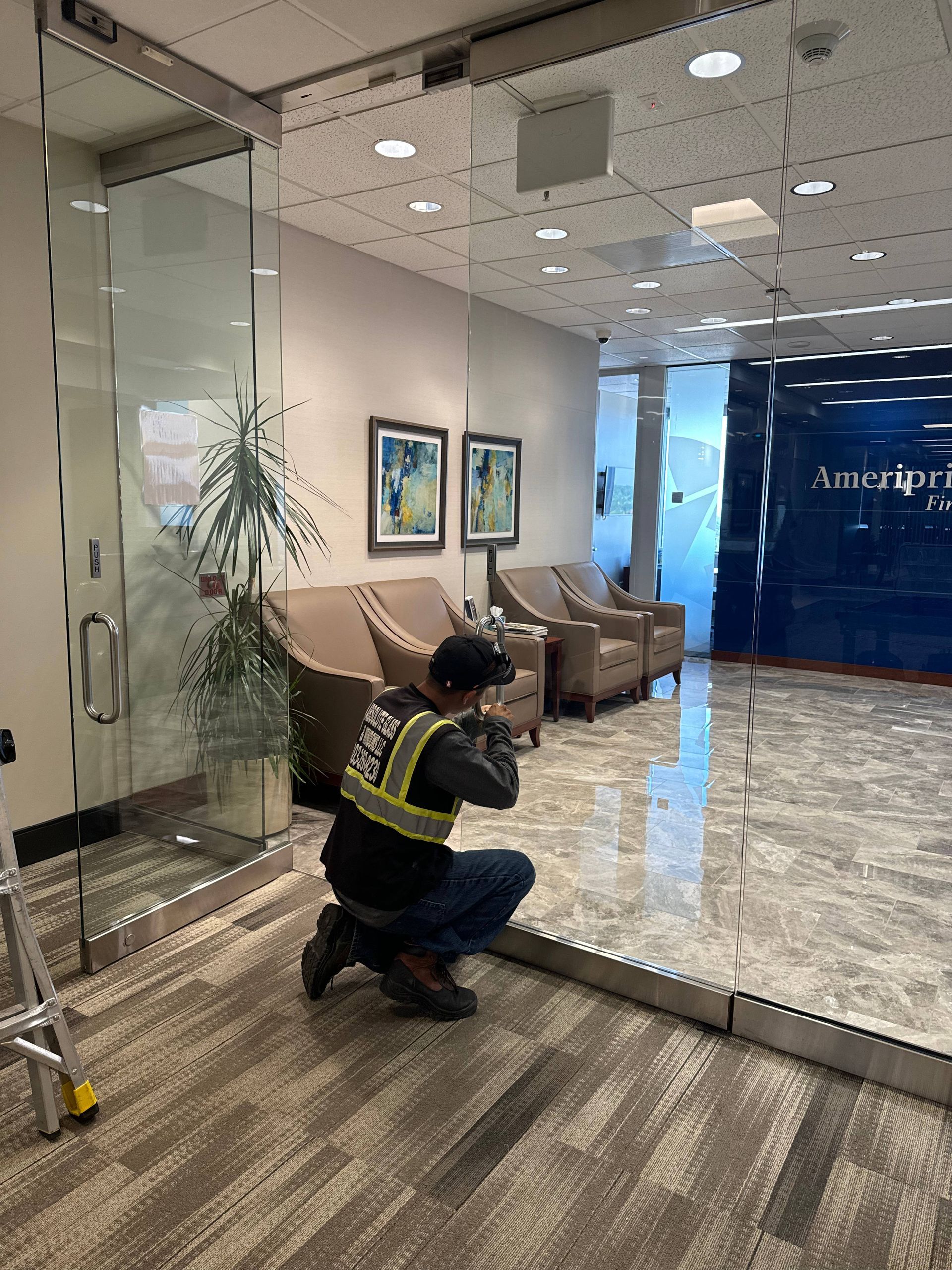 Person in work clothes kneels on floor, working on glass door in office lobby with furniture and an American Eagle sign.