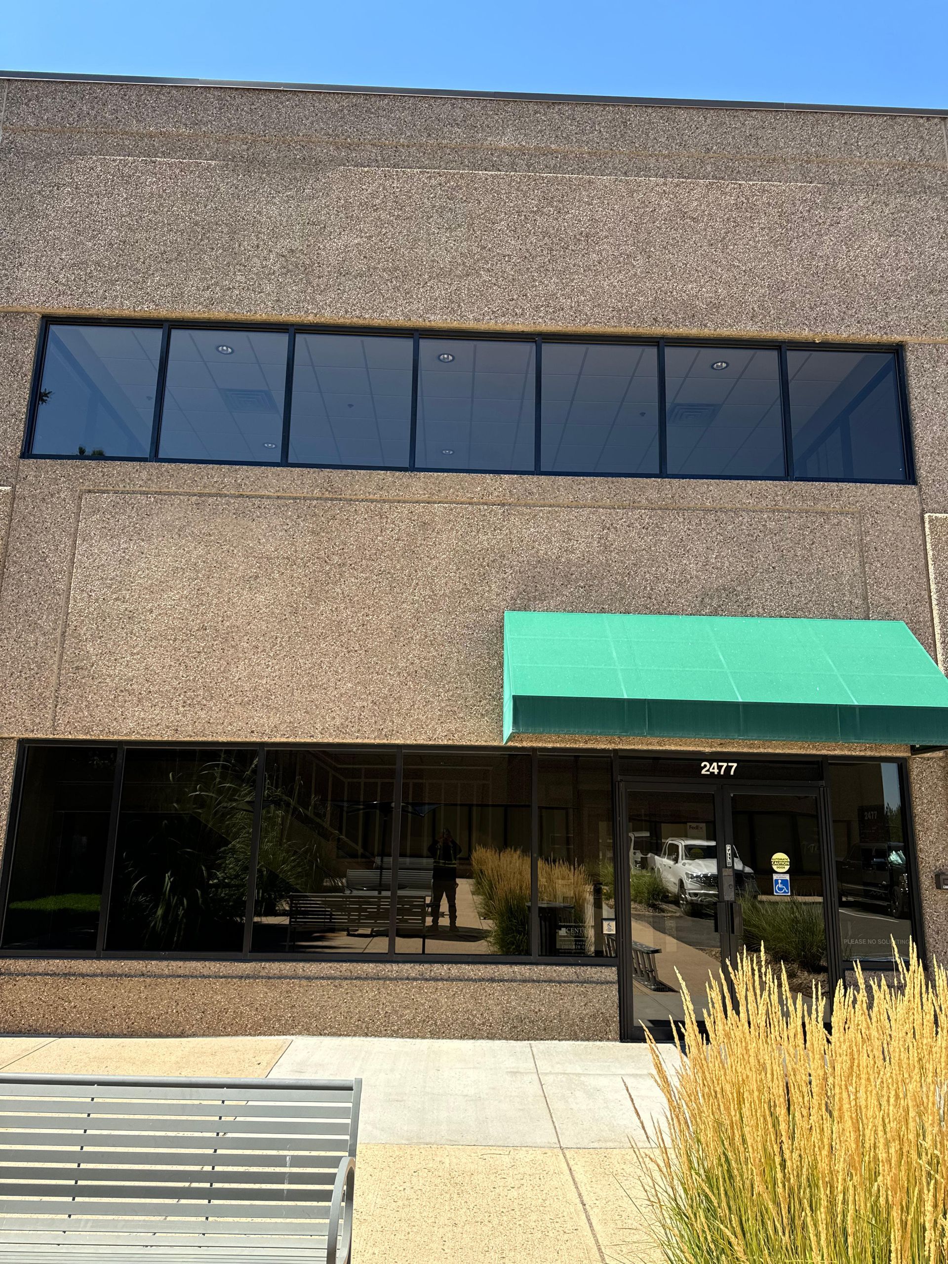 Exterior of a brick building with large windows. Green awning over the entrance, bench in front.