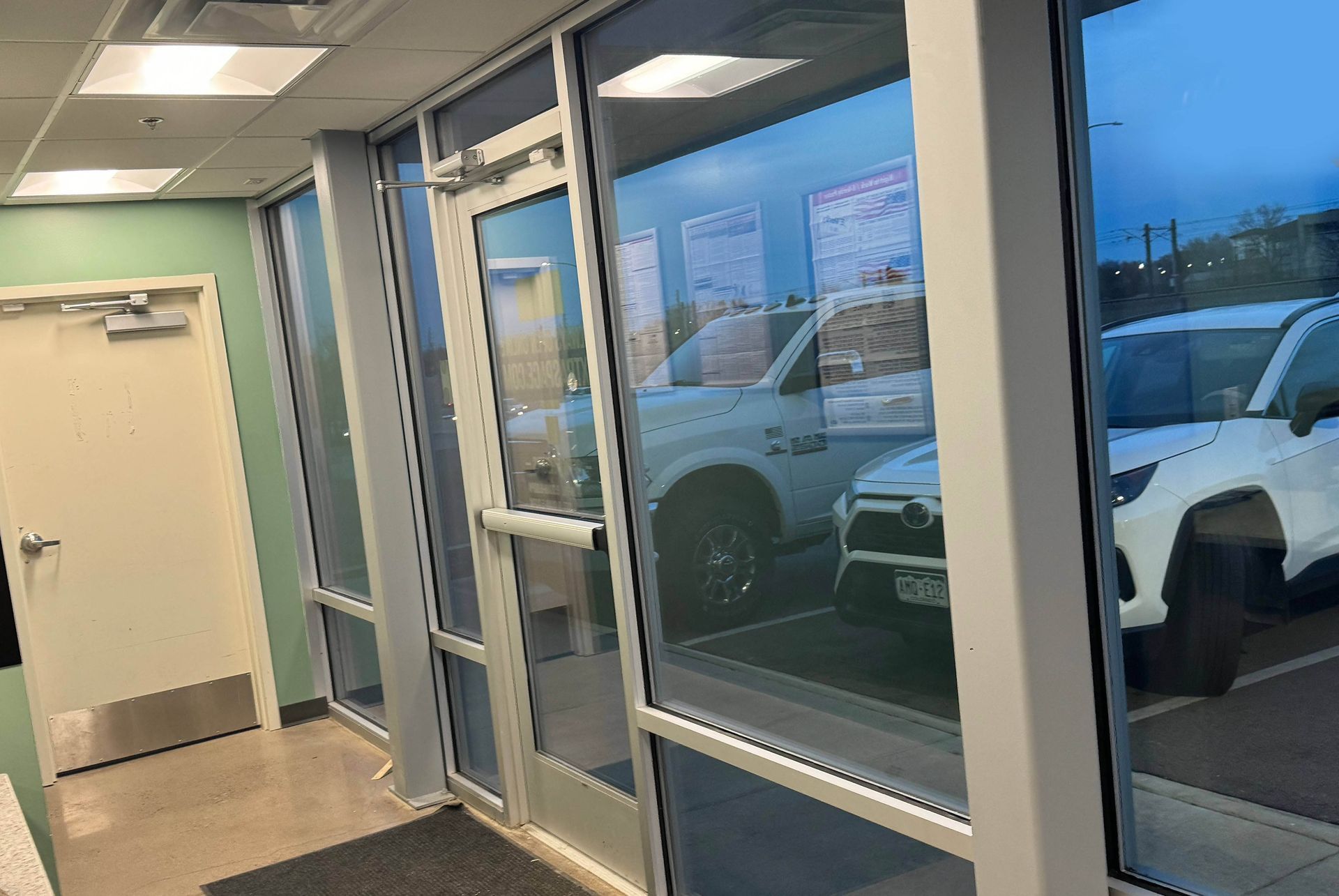Interior view of a building with glass windows and doors, reflecting parked cars outside.