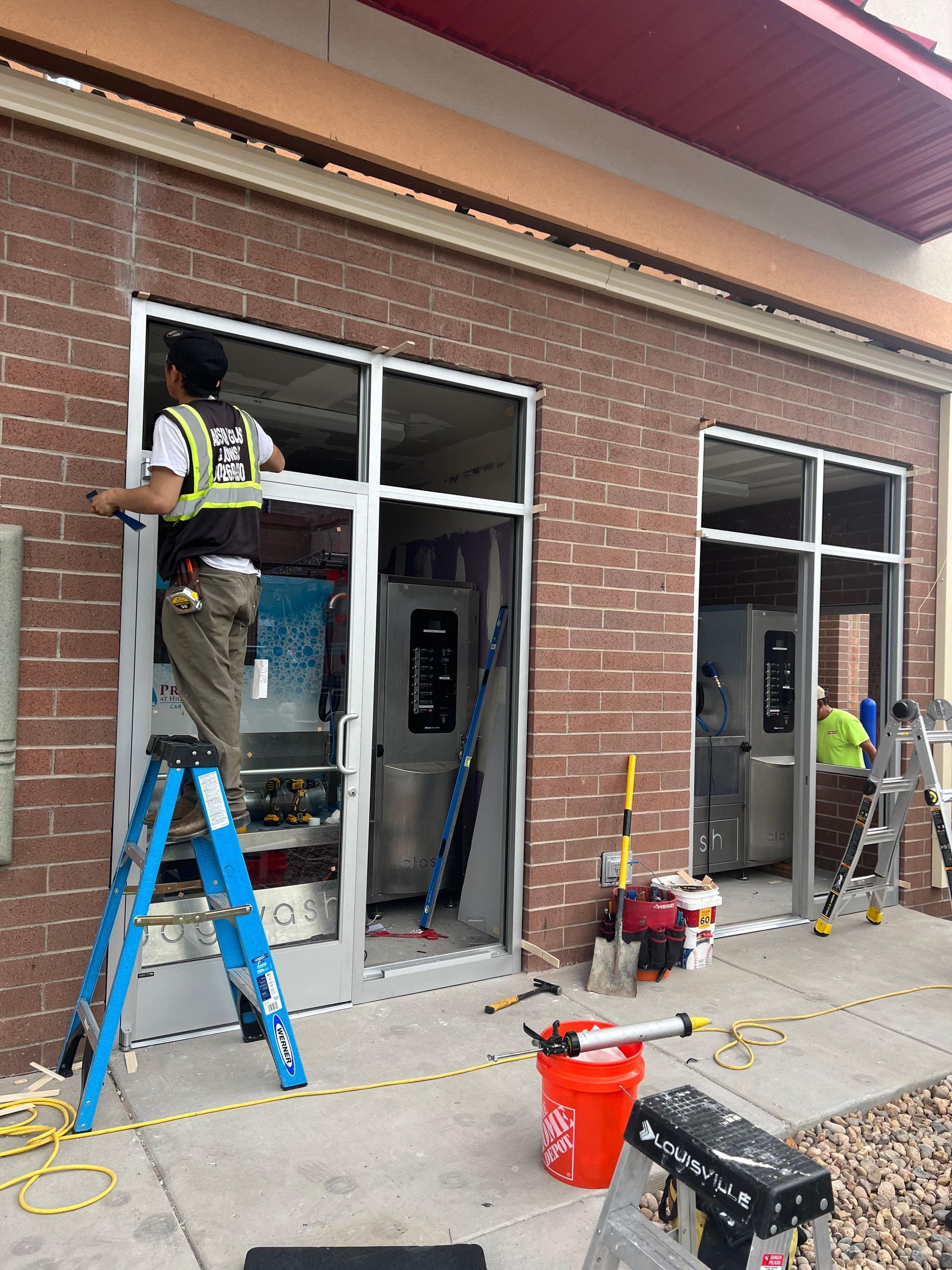 Construction workers install a storefront window on a brick building. One worker uses a ladder, others are nearby.