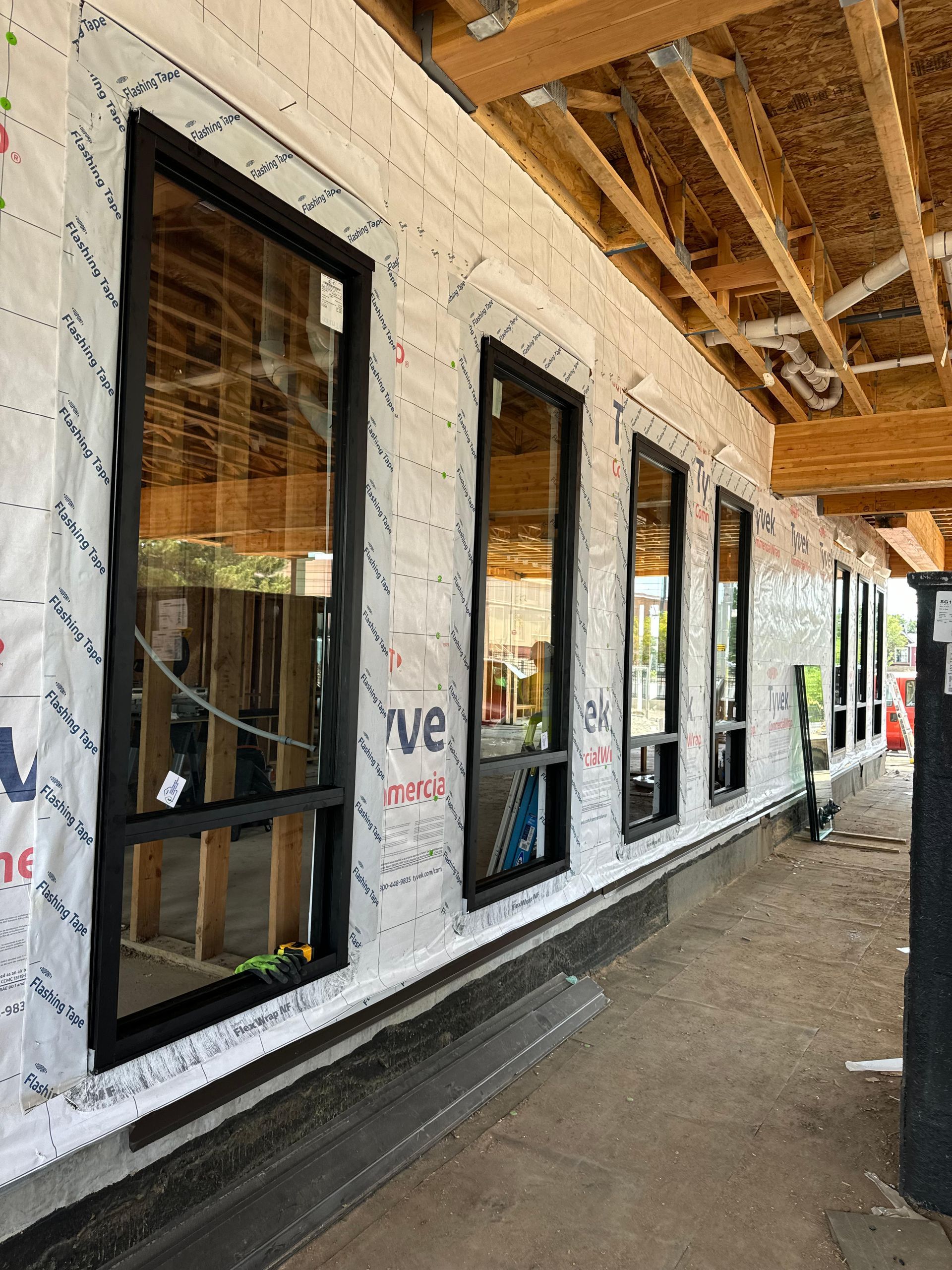 Exterior view of a building under construction with black-framed windows. The wall is covered with protective wrap.