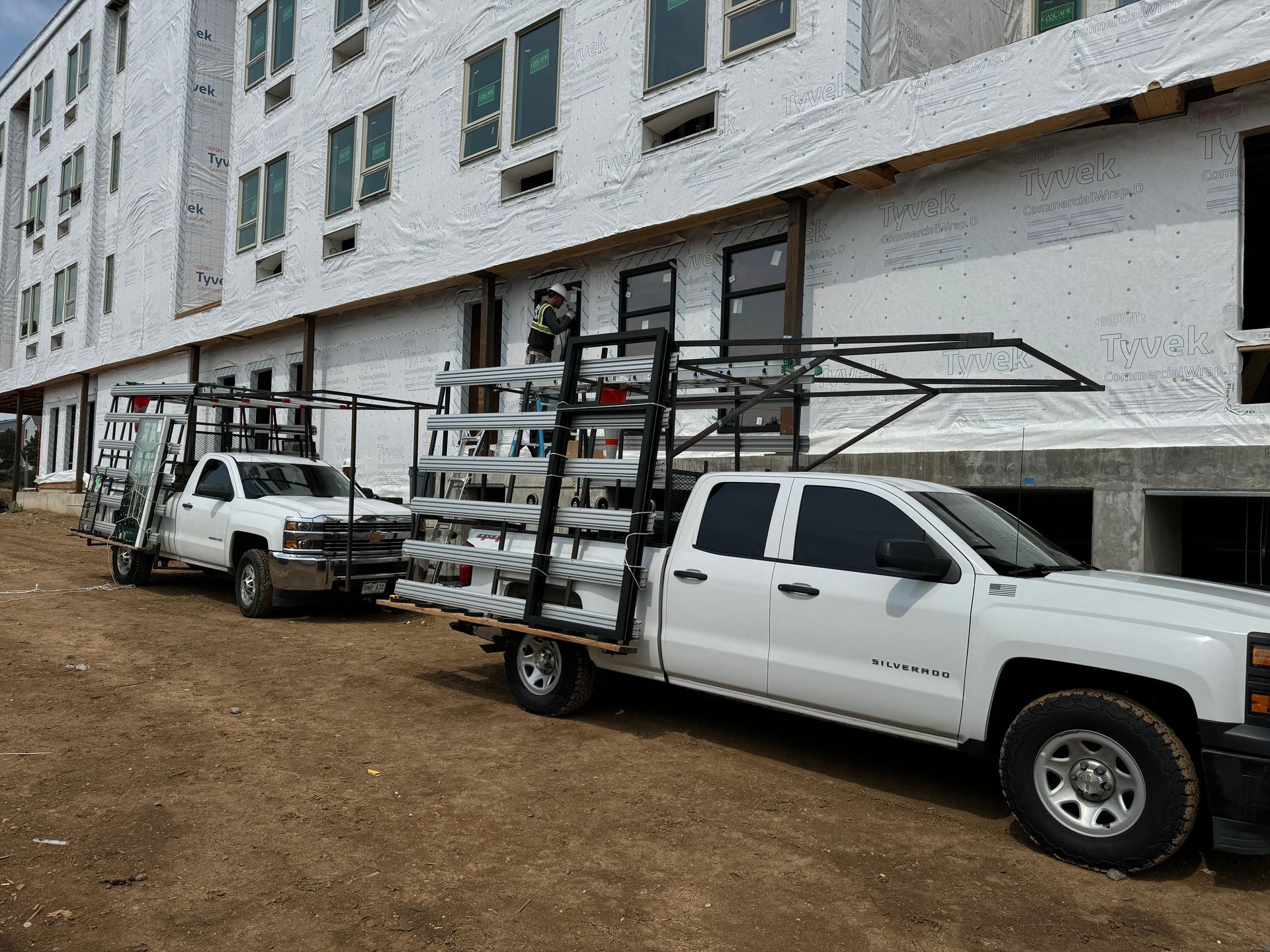 Two white trucks with racks of metal building materials at a construction site.