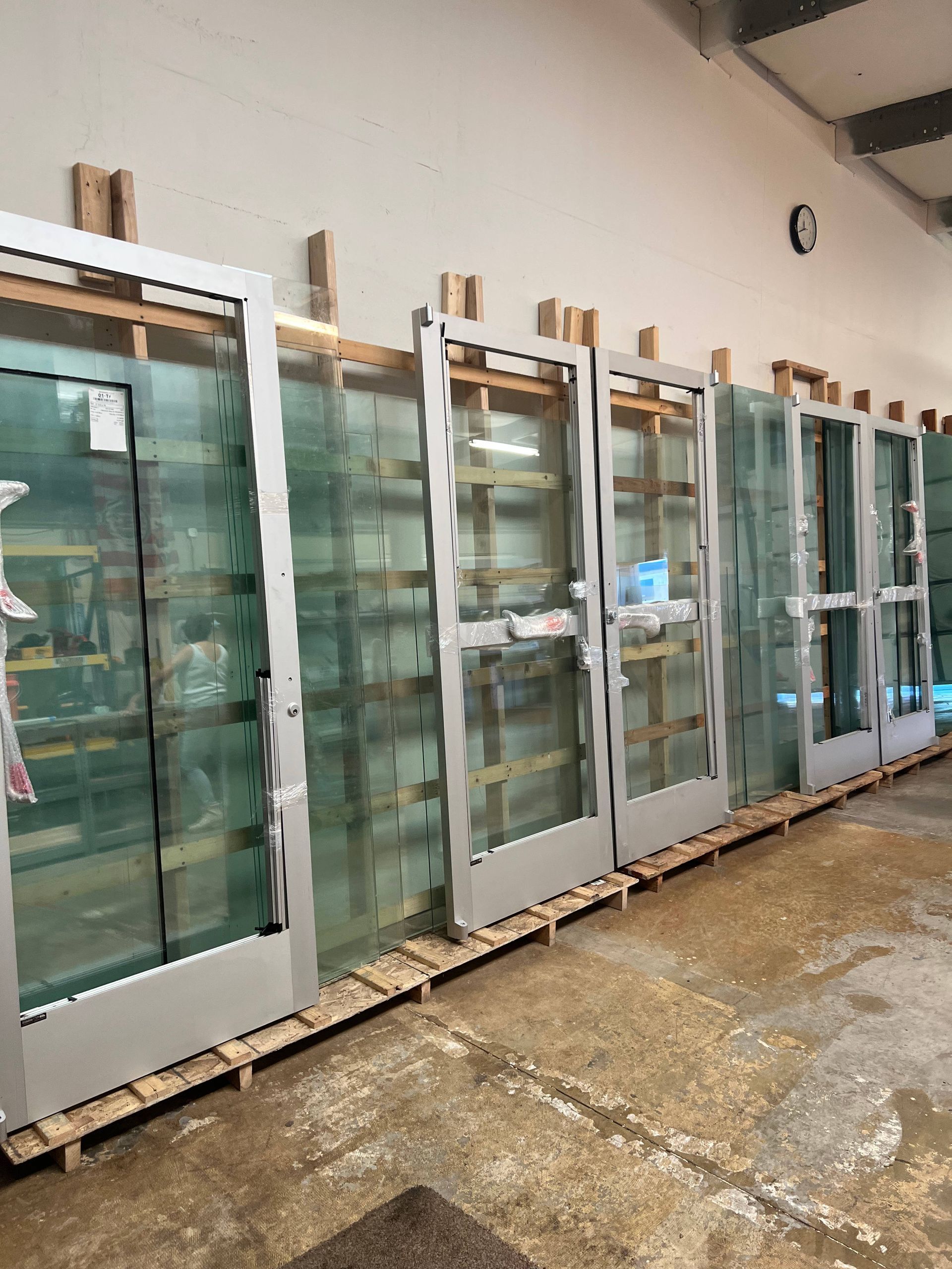 Glass doors in silver frames, leaning against a wall, stored on wooden pallets inside a warehouse.