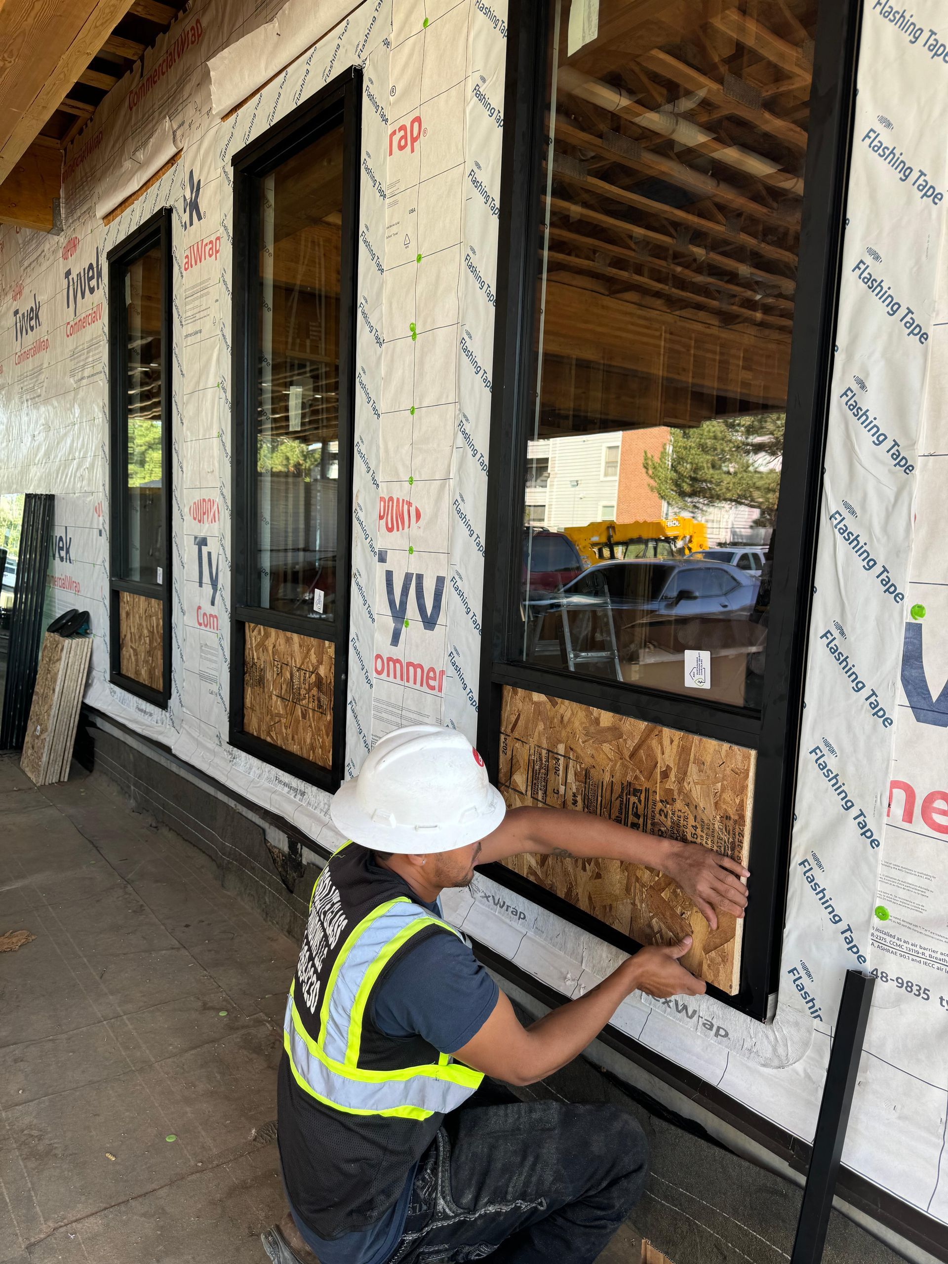 Construction worker installing wood panel beneath a large window with black frame on a building exterior.
