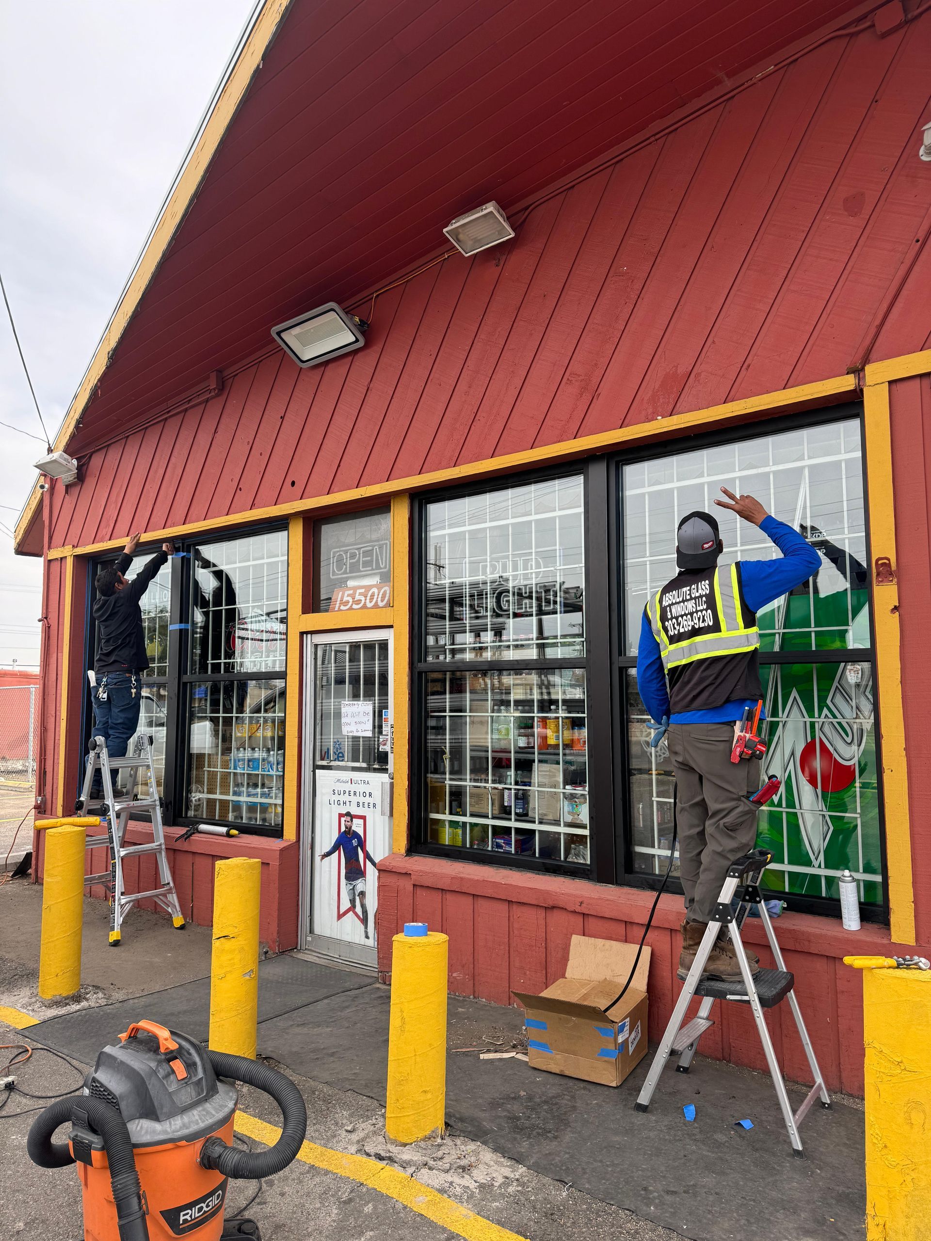 Workers cleaning windows of a red building, one on a ladder, wearing safety vests.