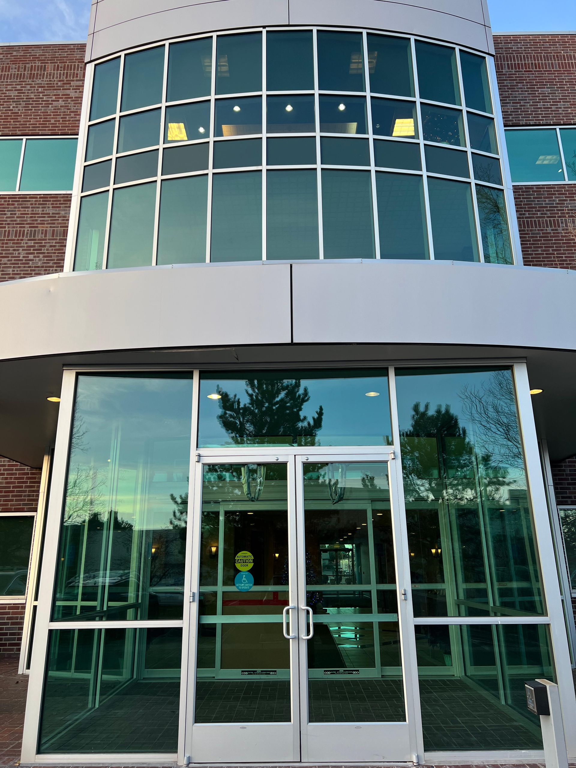 Glass doors of a modern building with curved glass windows above, reflecting trees and sky.