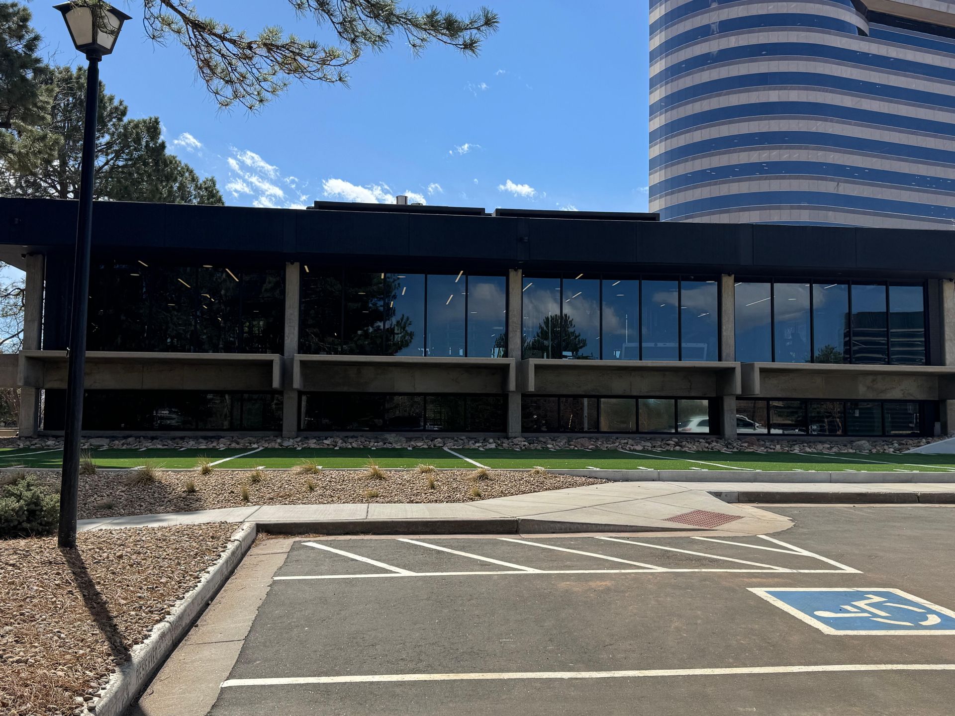 Building with large windows, sunny day. Parking spots and a tall blue-tinted building in the background.