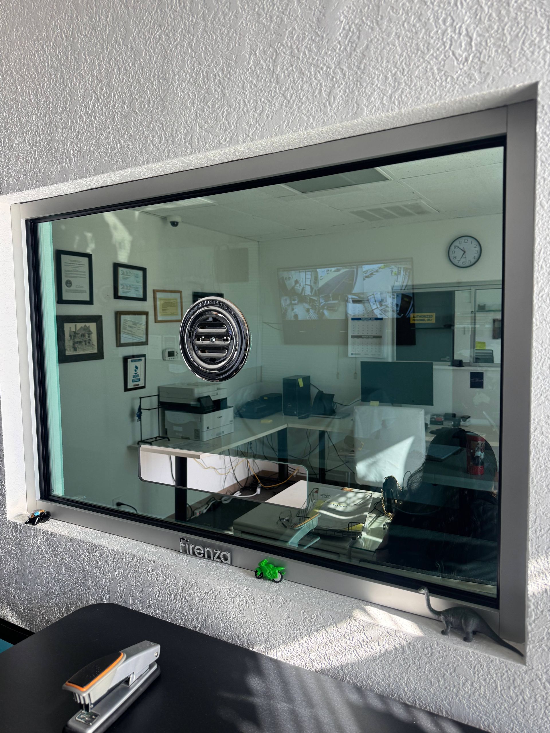 A silver-framed window showing an office with a desk, chairs, and a clock. A stapler and toy car sit on a table below.