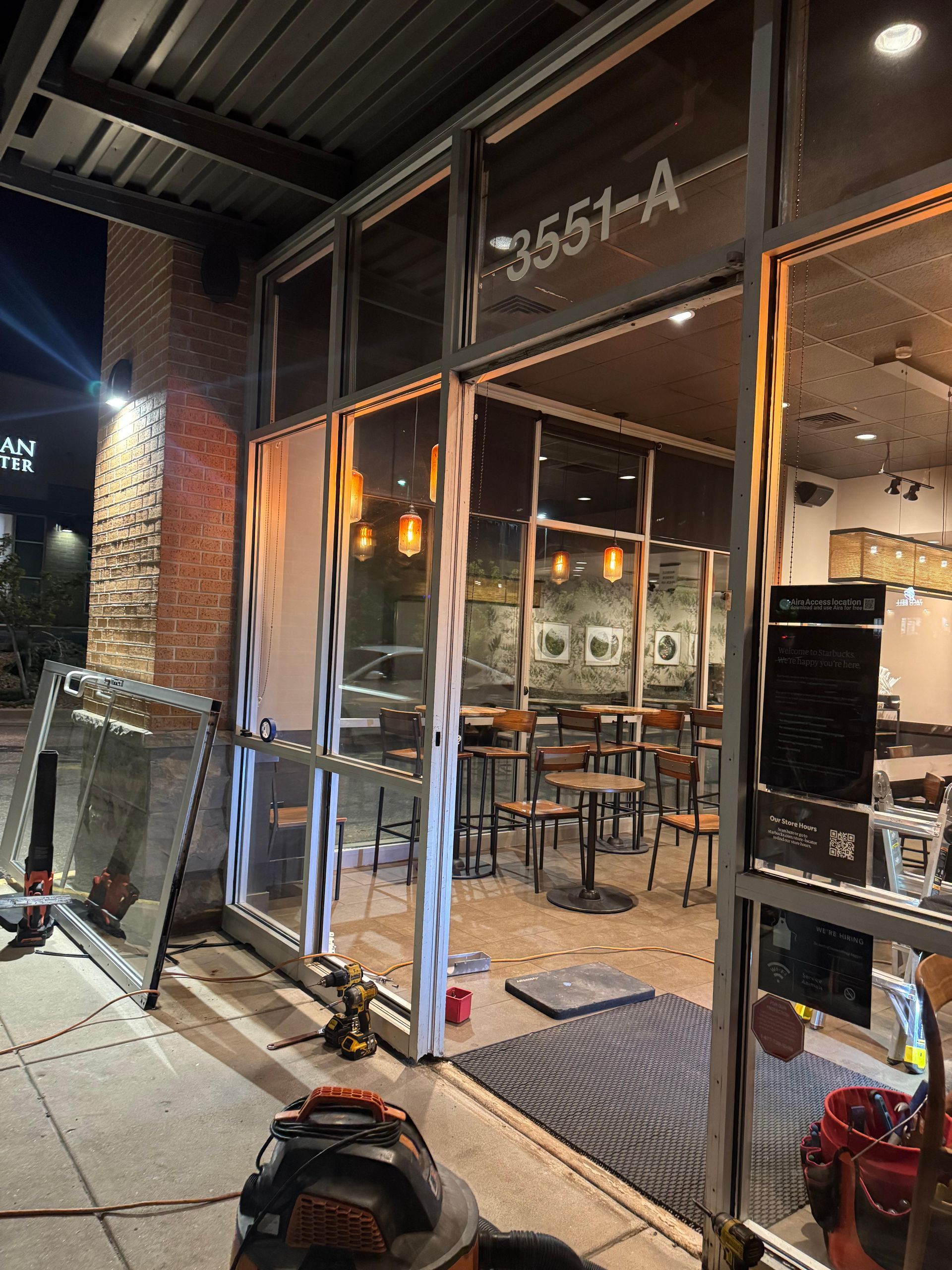 Broken storefront window; construction workers fixing it. Building exterior with tables and chairs visible inside.