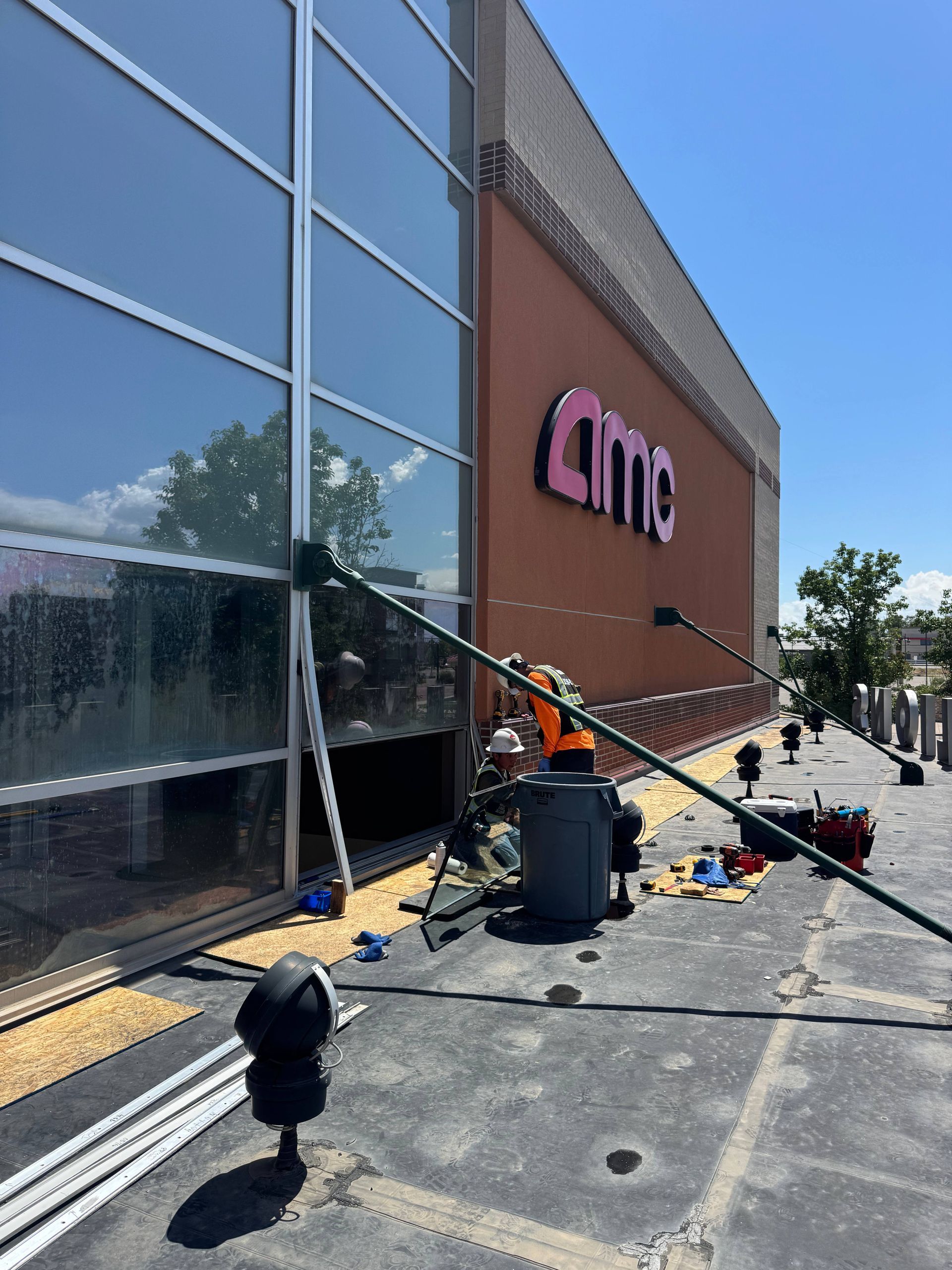 Workers on rooftop of AMC theater; installing equipment. Large windows and pink AMC logo visible. Blue sky.