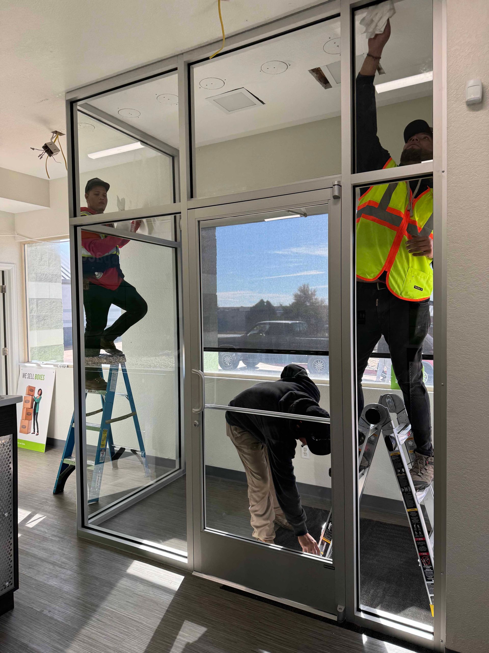 Workers installing a glass door and frame in a bright office. Three men on ladders, working with tools.