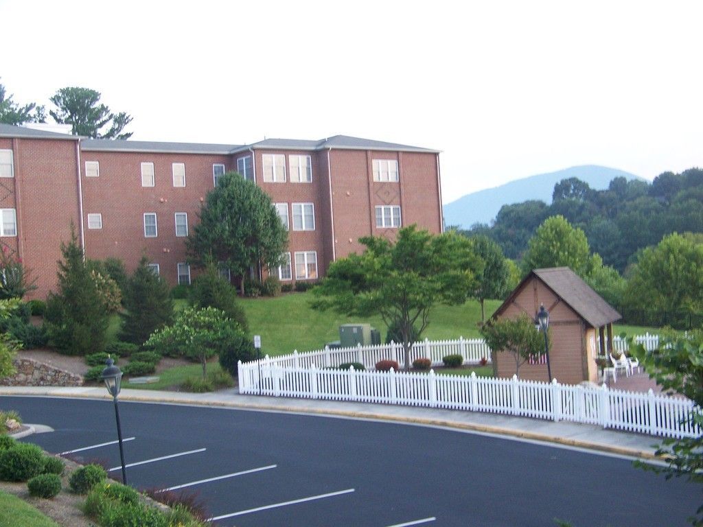 Brick apartment building next to a white picket fence, a small building and parking lot, with green trees and mountains.
