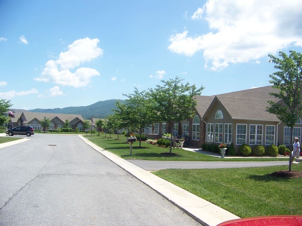 Paved street lined with single-story homes, green lawns, and trees under a blue sky, mountains in the distance.