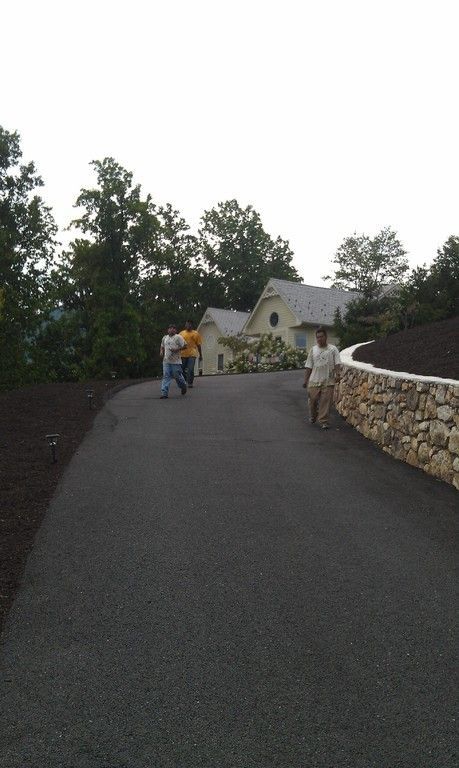 Asphalt driveway leads to a light-colored house. People walk on the driveway. A stone wall is on the right.