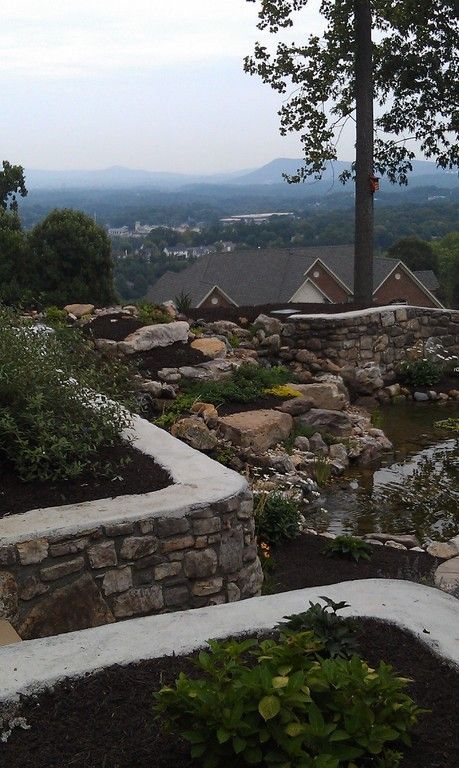 Stone retaining walls and garden with a view of a town nestled in a valley. Overcast sky.