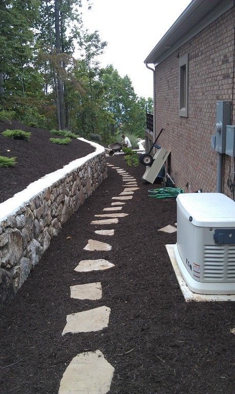 Stone path alongside a retaining wall with mulch and a generator next to a brick building.