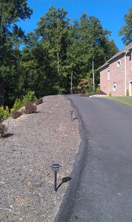 Asphalt driveway with solar lights, next to a landscaped slope with bushes. Trees and a brick building in the background.