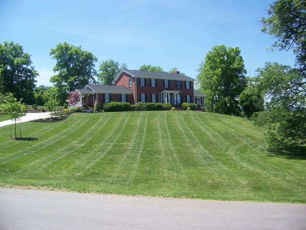 Red brick house on a grassy hill, striped lawn, trees, and a clear blue sky.