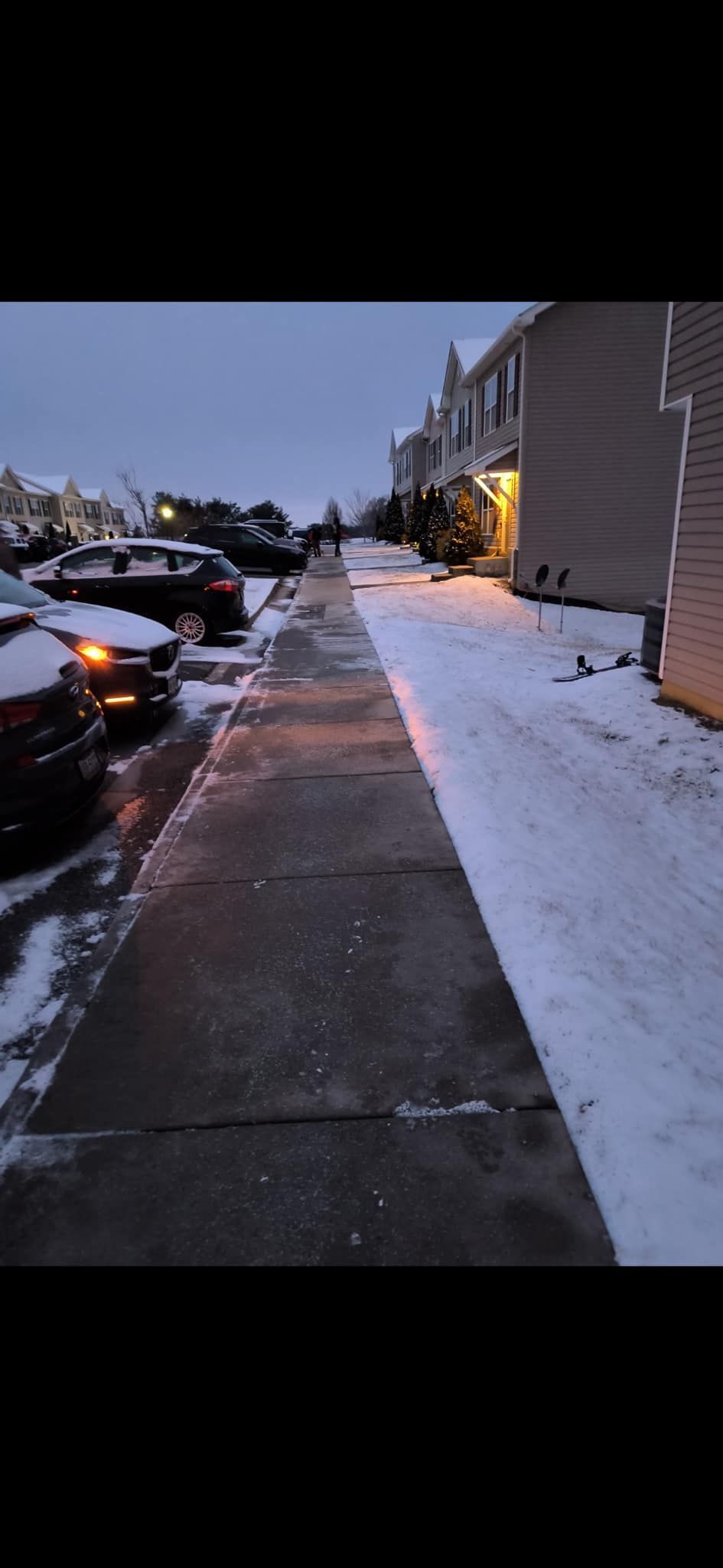 Snowy sidewalk lined with parked cars and townhouses under a dim sky.