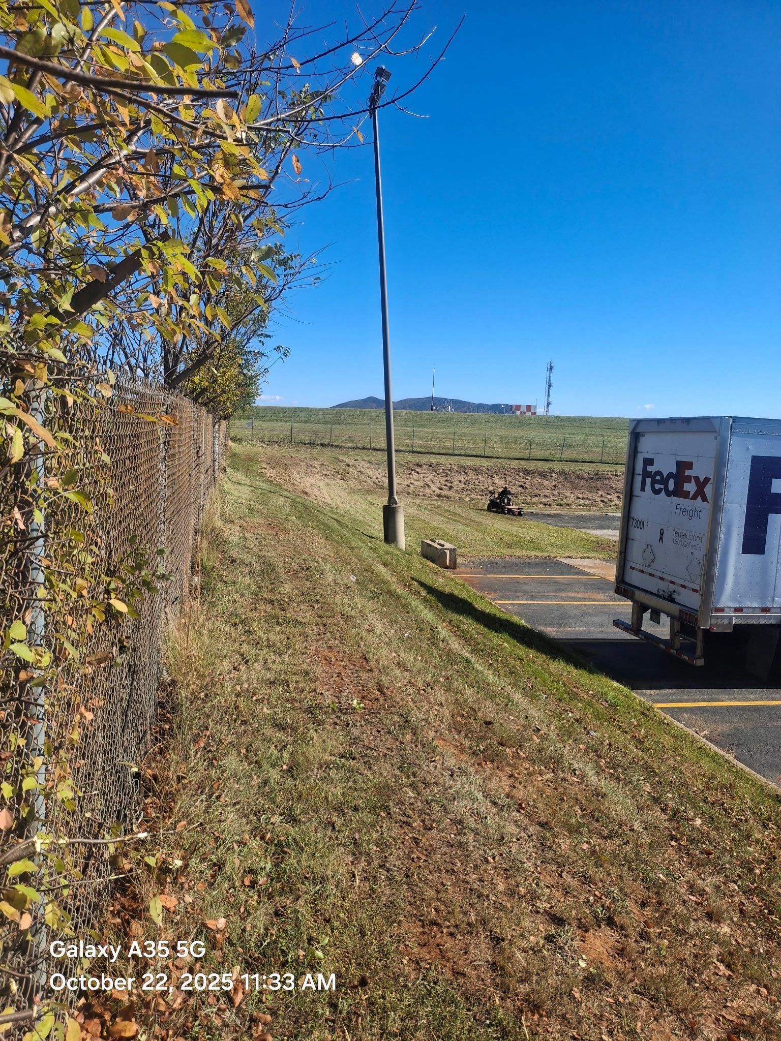 A FedEx truck parked near a chain-link fence and a grassy embankment under a blue sky.