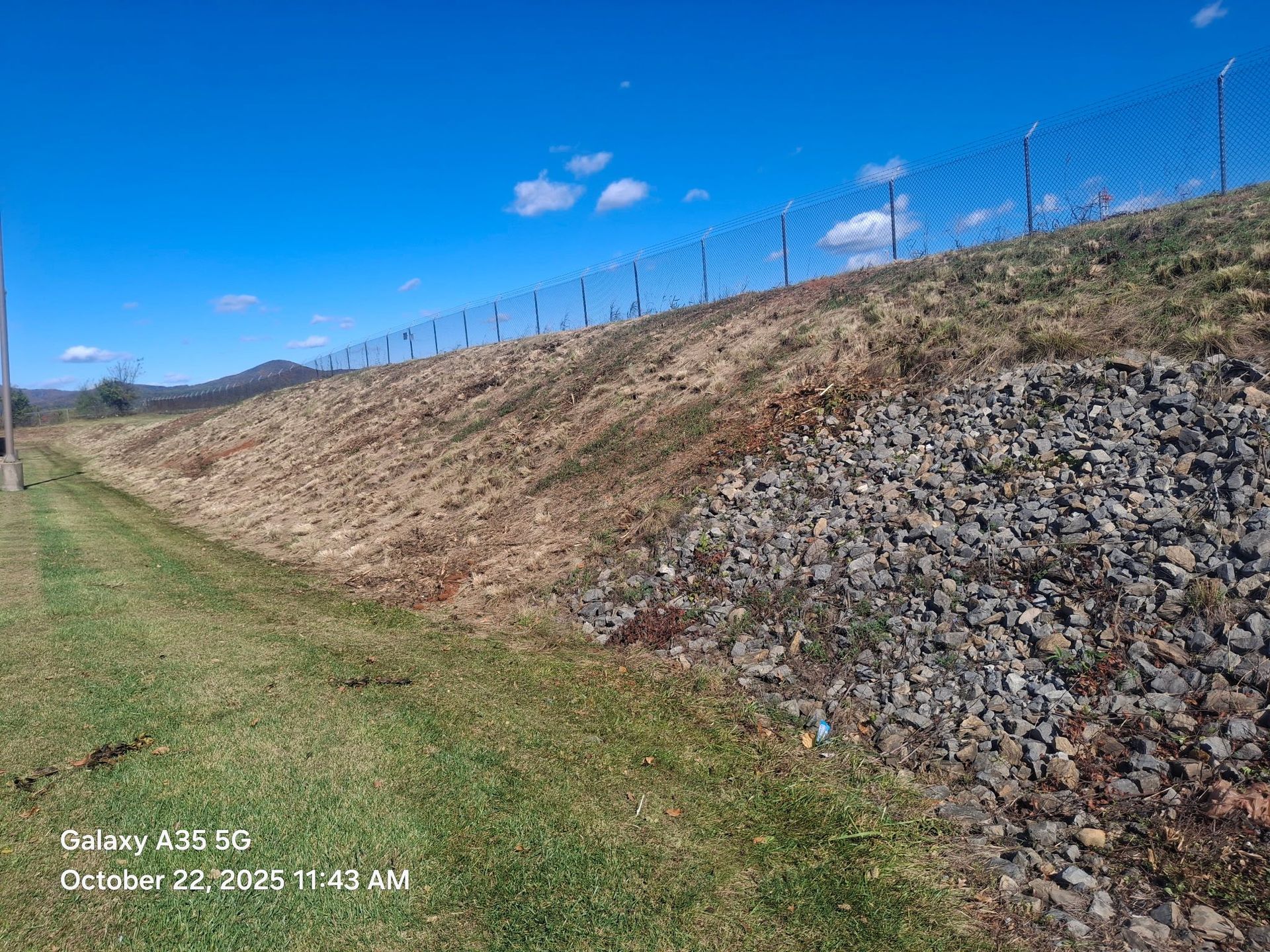 A chain link fence atop a grassy embankment lined with rocks, under a blue sky.