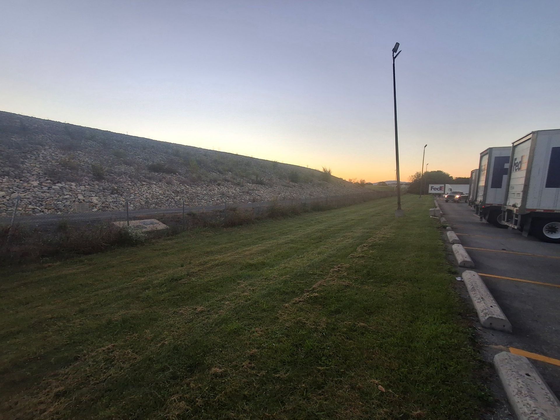 Grassy area next to a rock-covered embankment and parked semi-trucks under a dusky sky.
