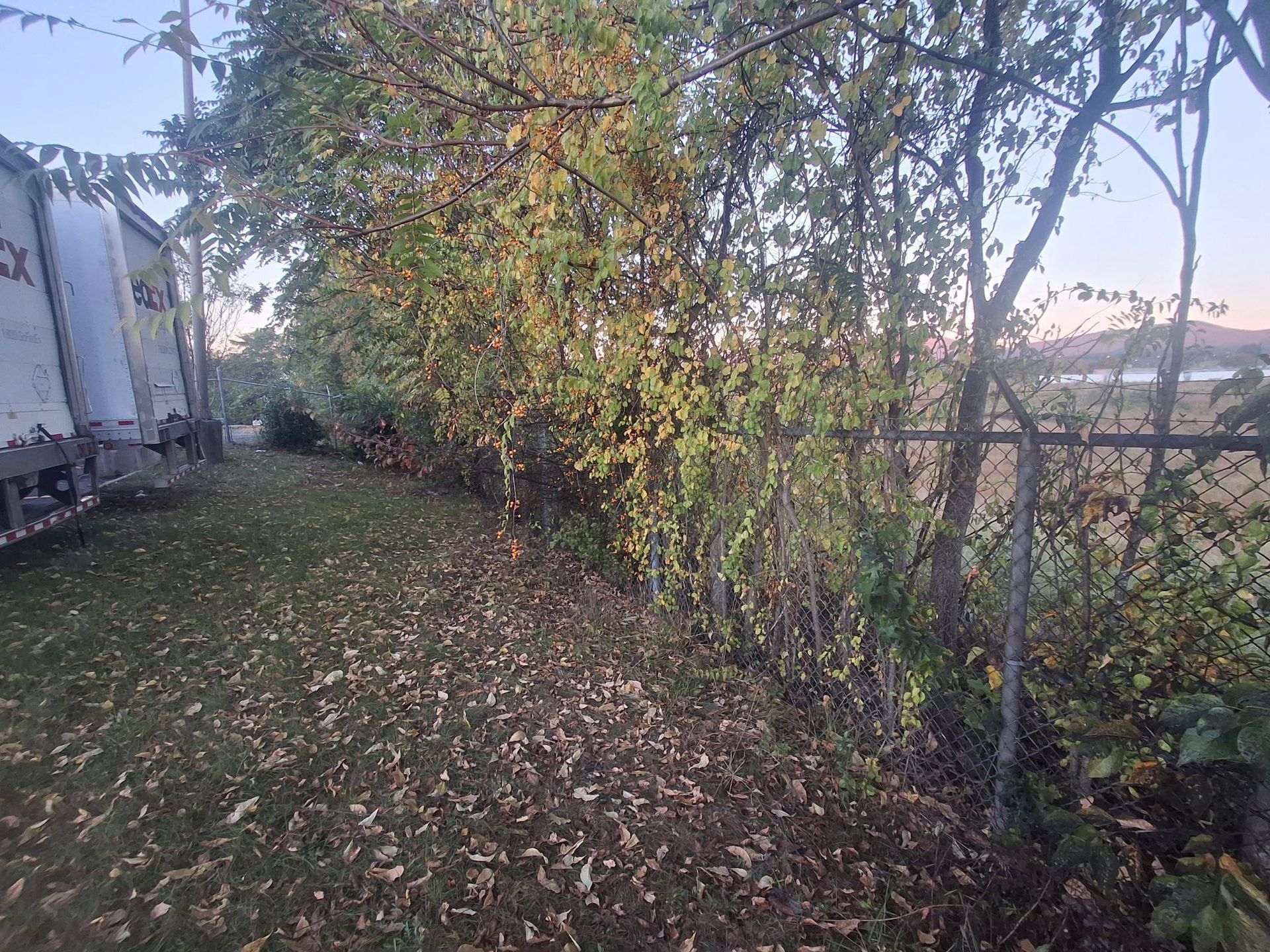 Path through a wooded area next to a chain-link fence and parked truck trailers; foliage with some autumn colors.