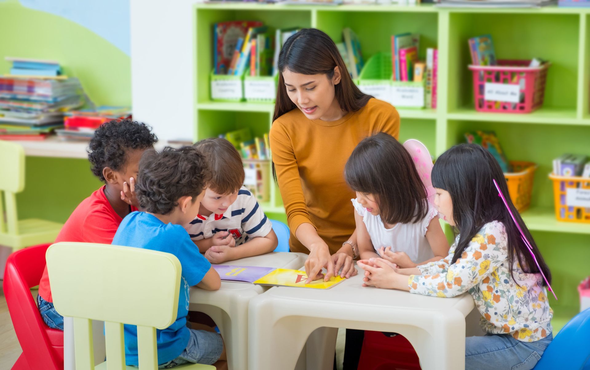 Teacher showing students a book at a table in a brightly colored classroom.