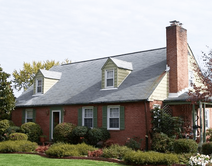 Brick suburban house with gray roof, dormer windows, chimney, and landscaped front yard