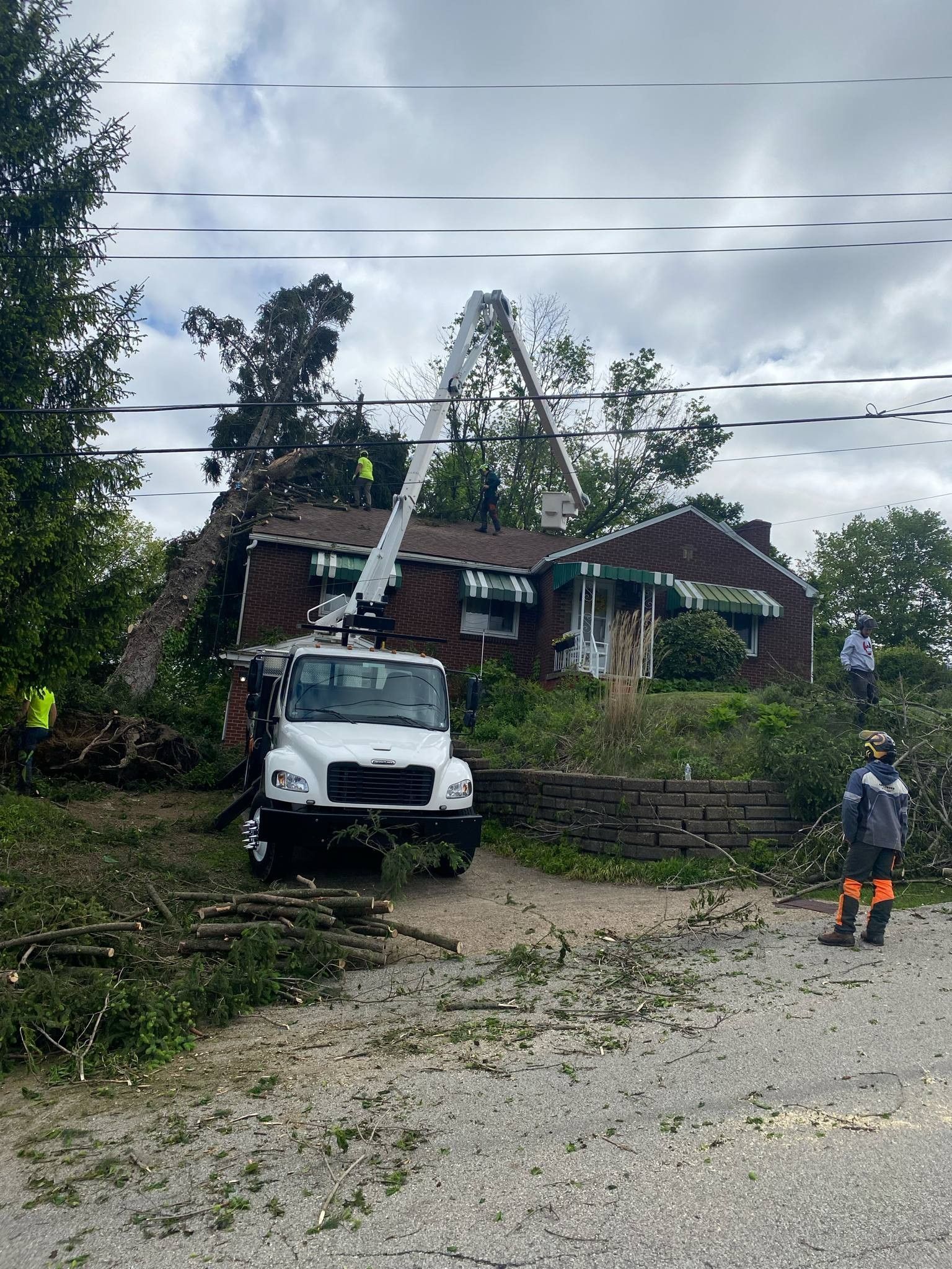 Tree removal service: Truck with lift cutting tree from house roof. Workers wear safety vests. Cloudy sky.