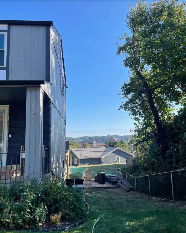 Two-story gray house next to a grassy yard and trees, with buildings visible in the distance under a blue sky.