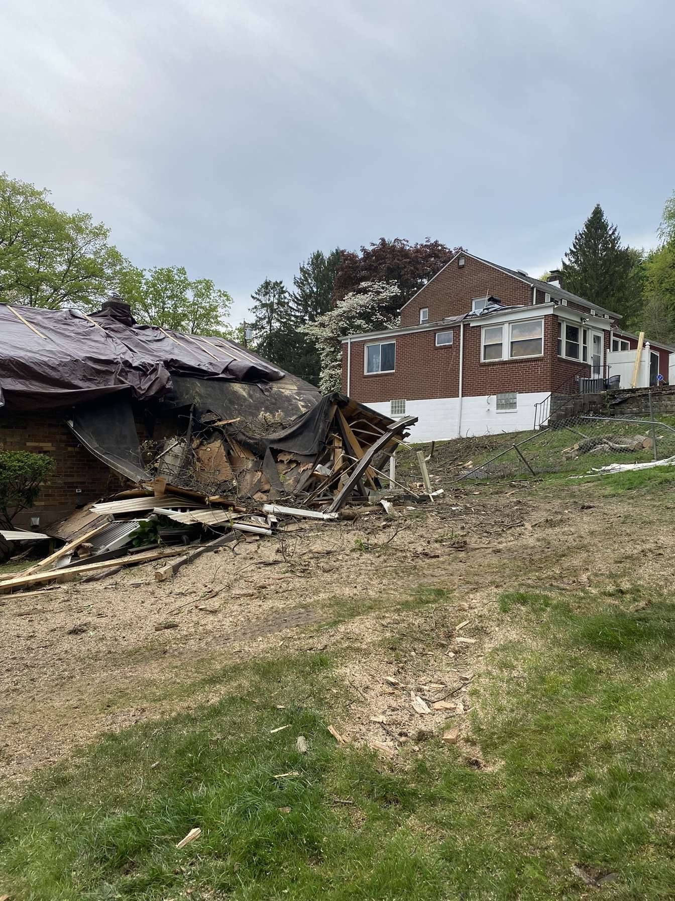 Two damaged houses on a grassy hillside, debris scattered. Cloudy sky overhead.