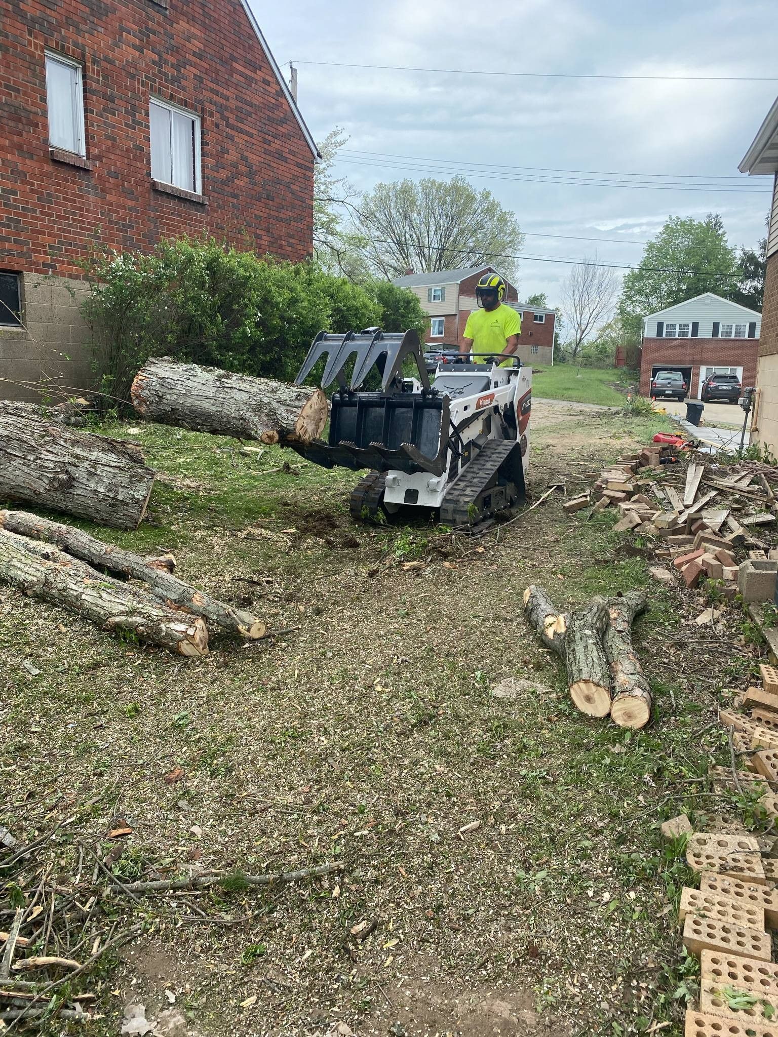 A worker uses a wood chipper on a mini skid steer to process logs near a brick building.