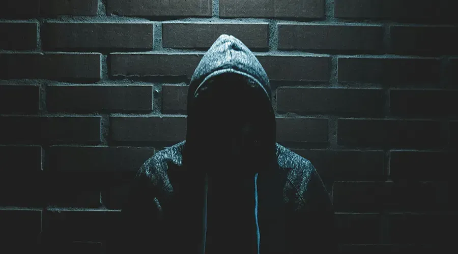 Hooded figure in shadow against a dark brick wall, lit by dim blue light