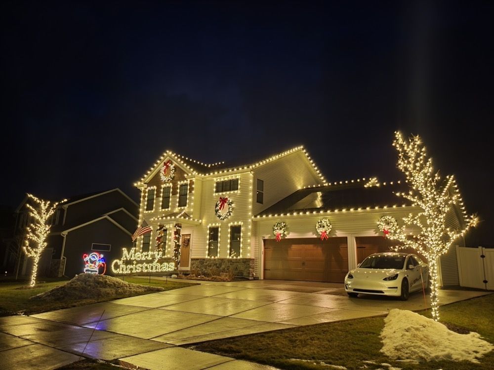 House decorated with warm white Christmas lights, glowing trees, and a festive holiday sign at night
