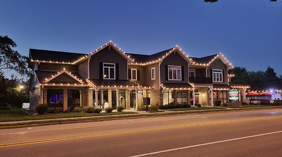 Small town storefronts at dusk, outlined with white holiday lights along a quiet street.
