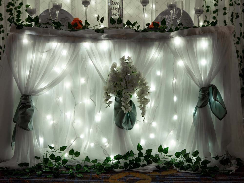 White draped backdrop with warm string lights, floral centerpiece, and greenery garland for a wedding or event backdrop