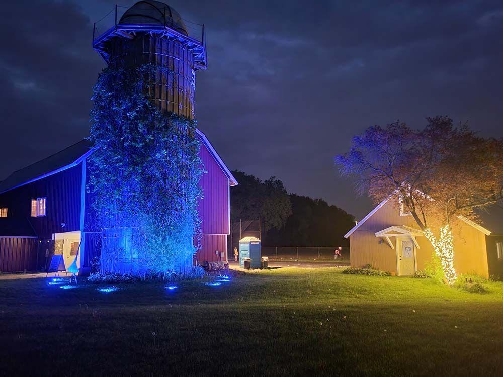 Nighttime outdoor scene with a blue-lit tower and a yellow-lit barn on a grassy field.