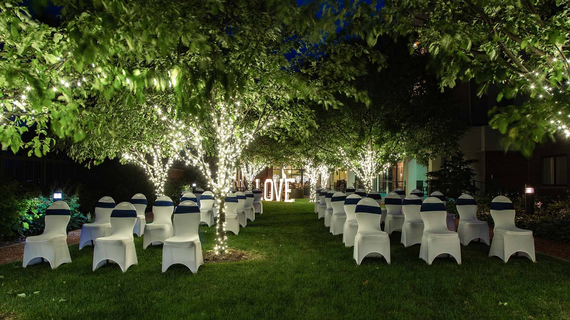 Outdoor wedding reception setup with white chairs around tables under trees lit by string lights at night