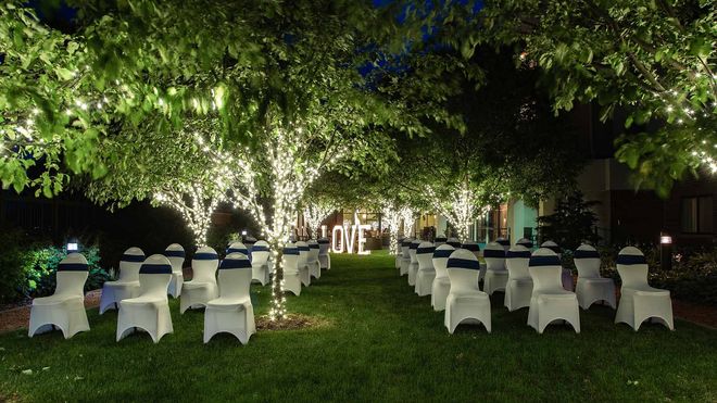 Outdoor wedding reception setup with white chairs around tables under trees lit by string lights at night