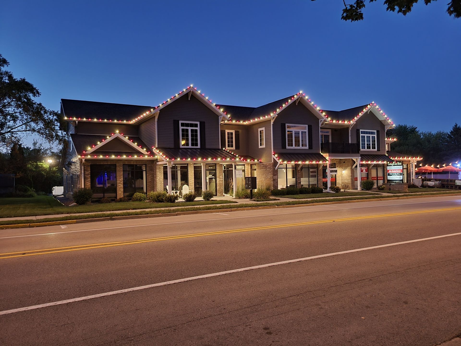 Street view of a lit row of townhouses at dusk, with holiday lights along the roofline.