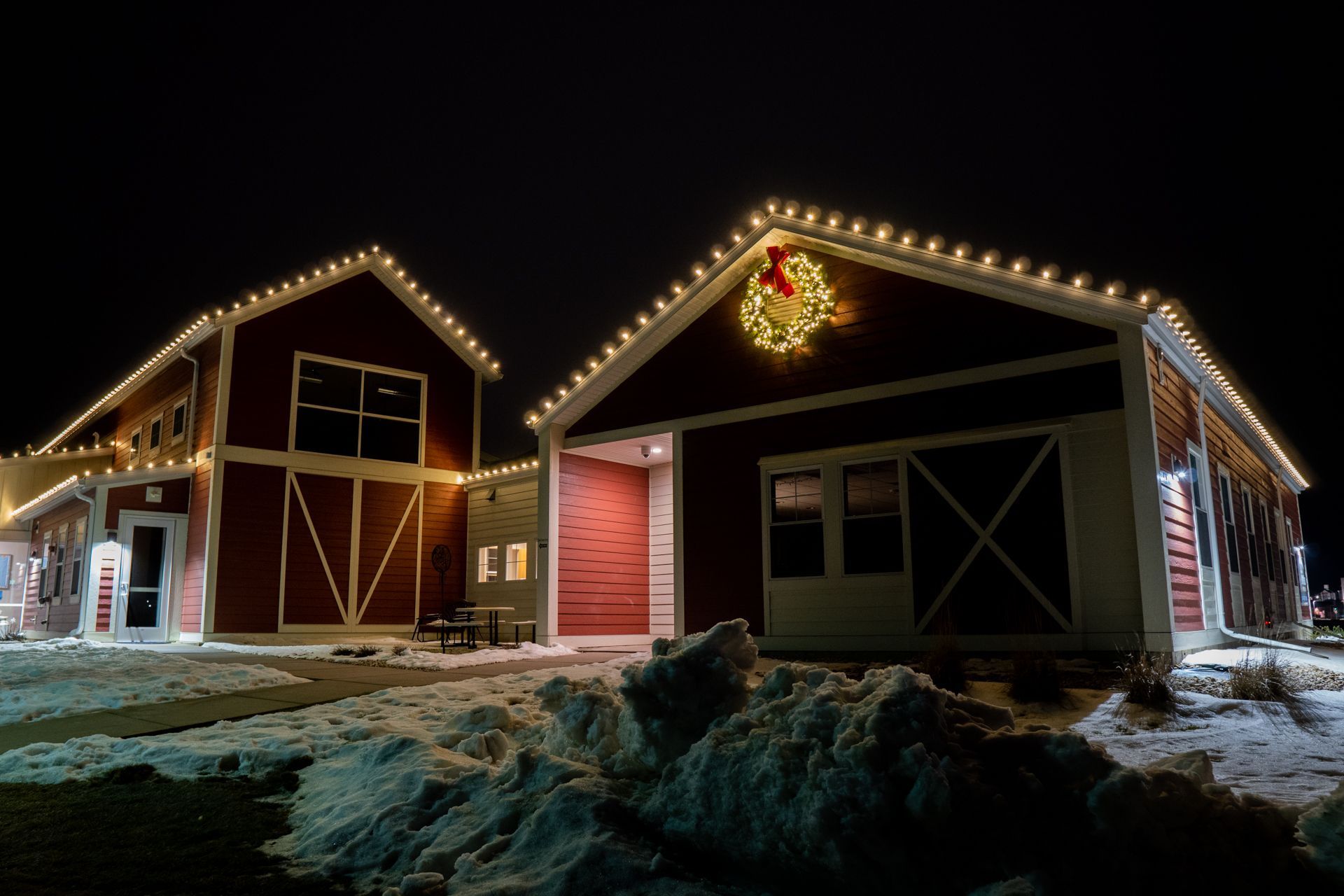 Snowy barn buildings outlined in warm lights at night, with a festive wreath on one roof.