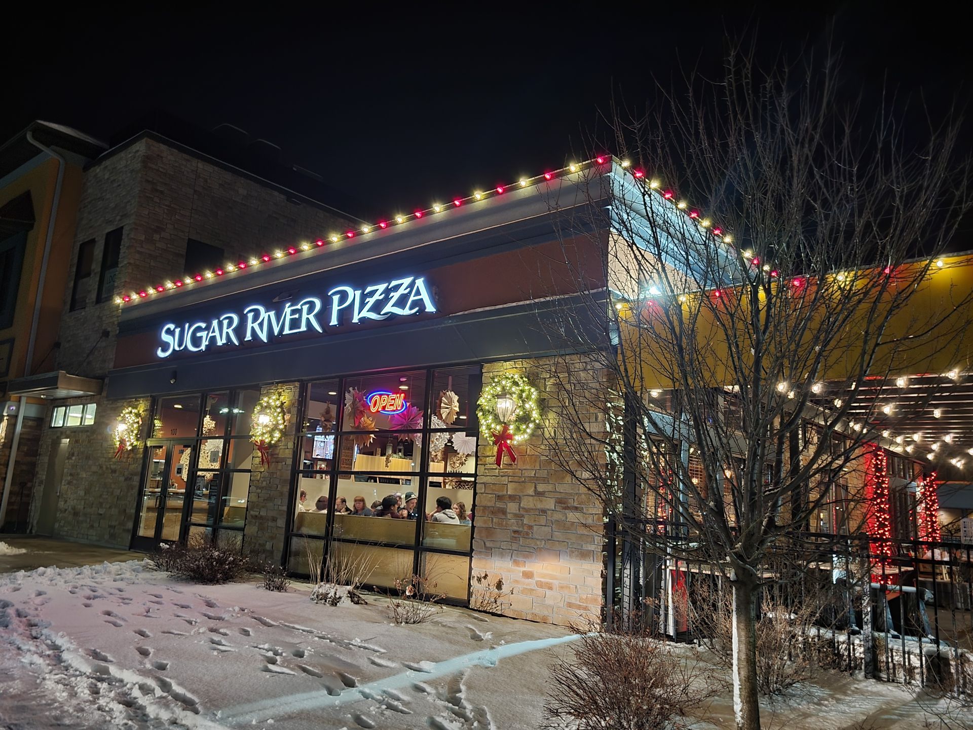 Sugar River Pizza storefront at night, lit with colorful holiday lights and snowy ground