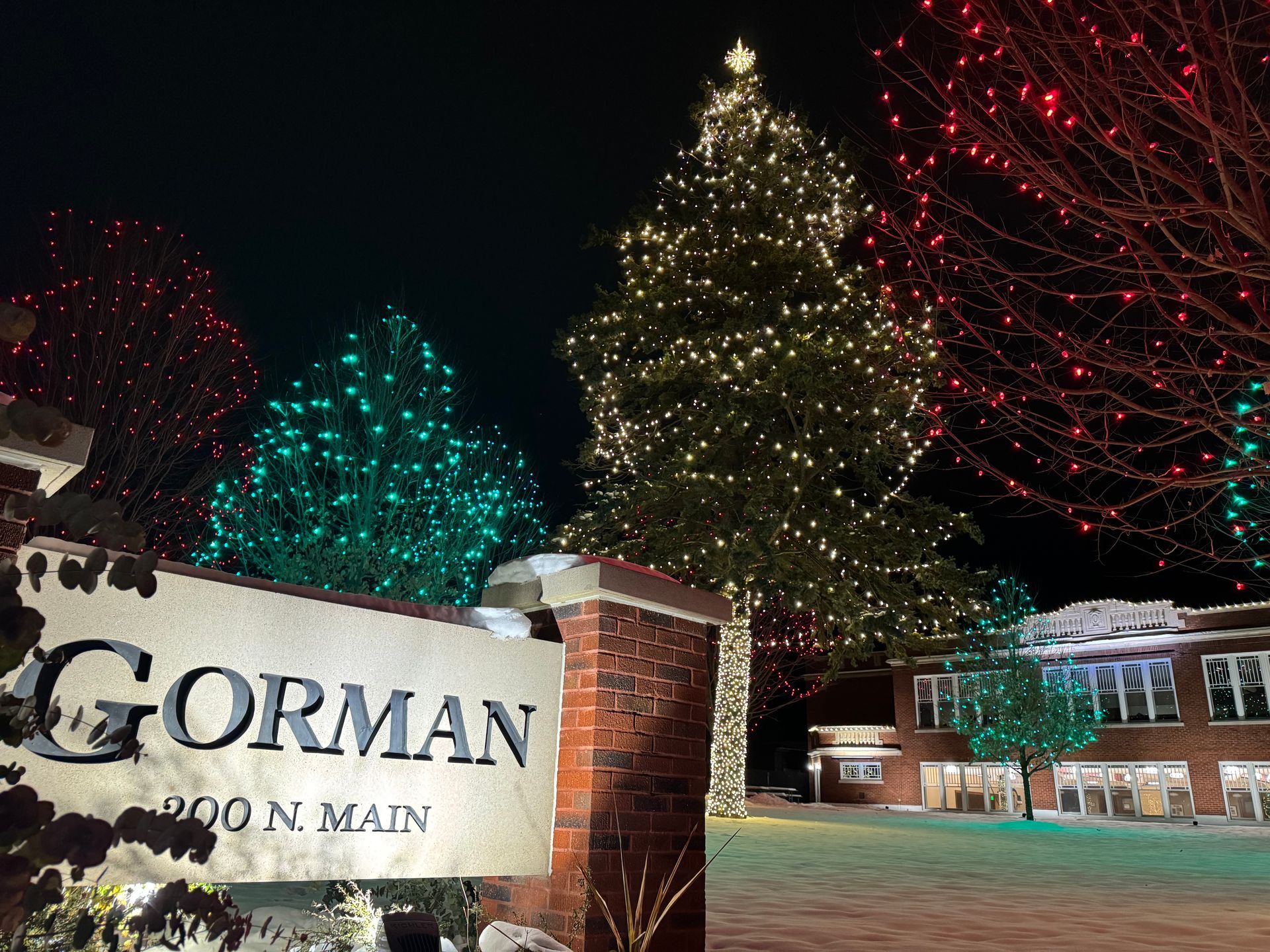 Gorman sign at night with illuminated Christmas trees and holiday lights outside a building
