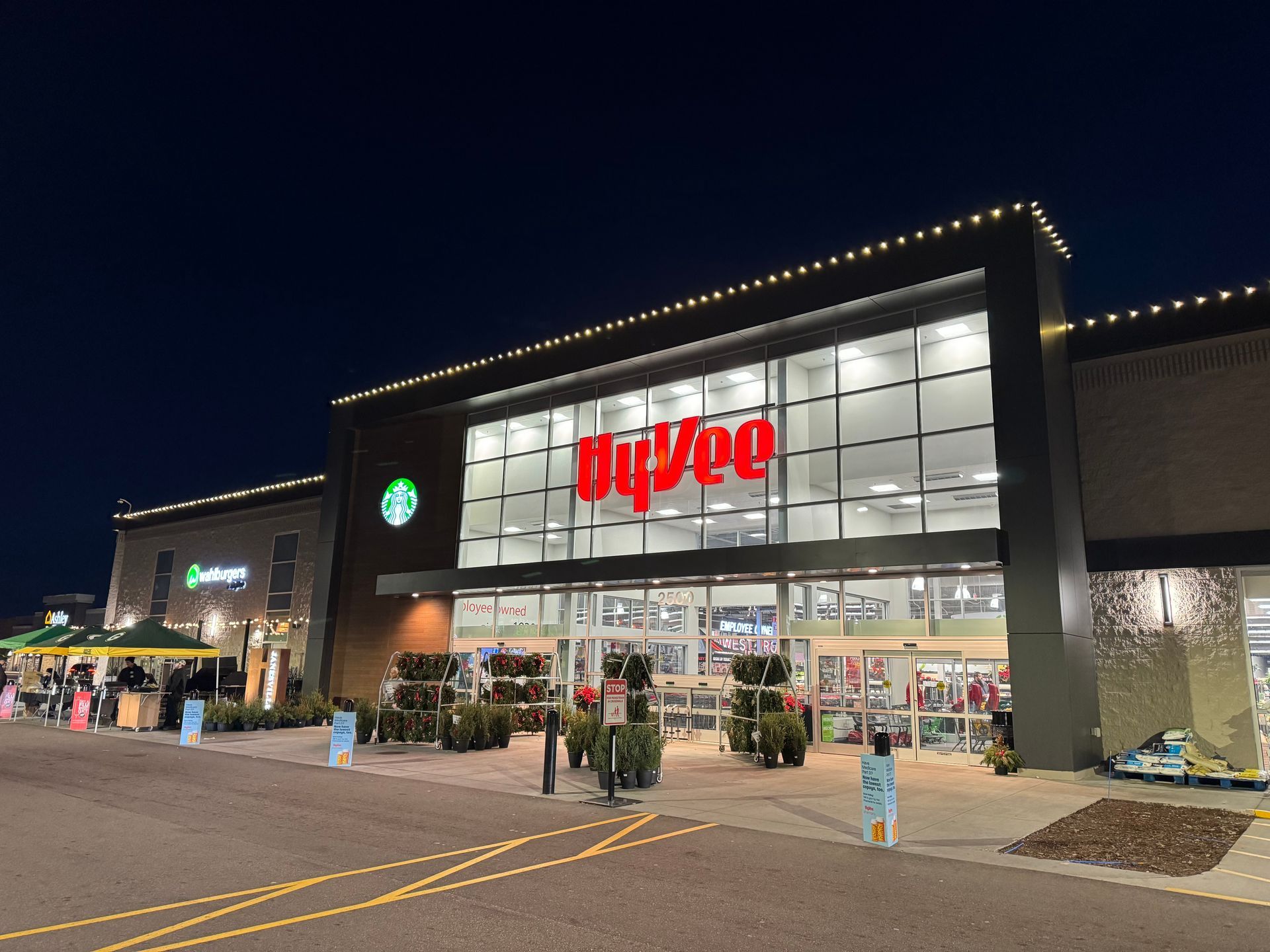 Nighttime exterior of a brightly lit grocery store with shoppers entering through glass doors.